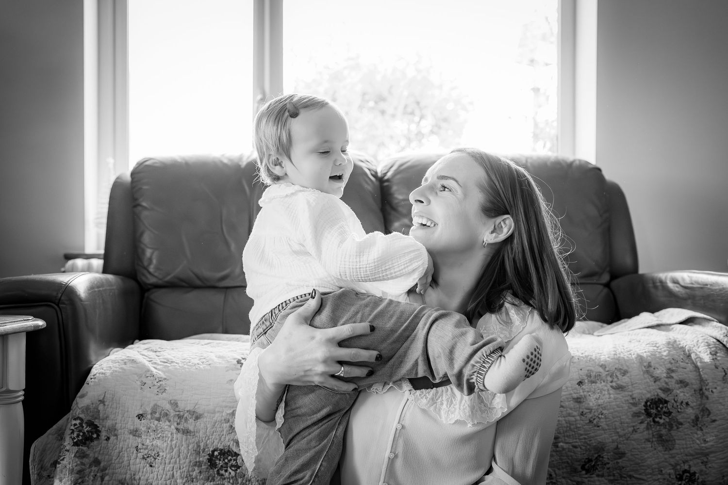 Mother and daughter in home session with yellow walls and beautiful window to back light them smiling and laughing. Black and White image.