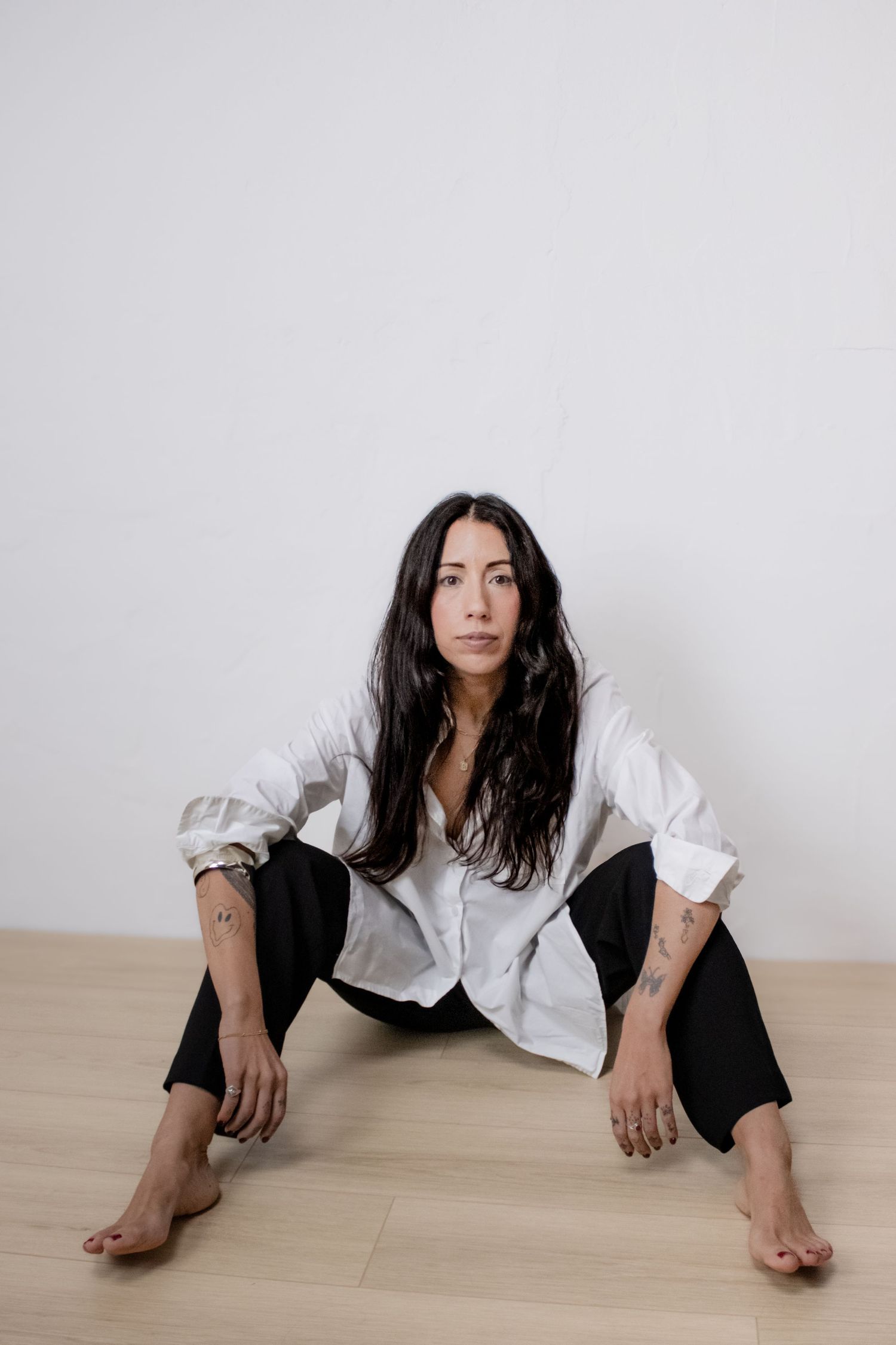 A person with long dark hair sits on wooden floor wearing an oversized white button-down shirt and black pants.