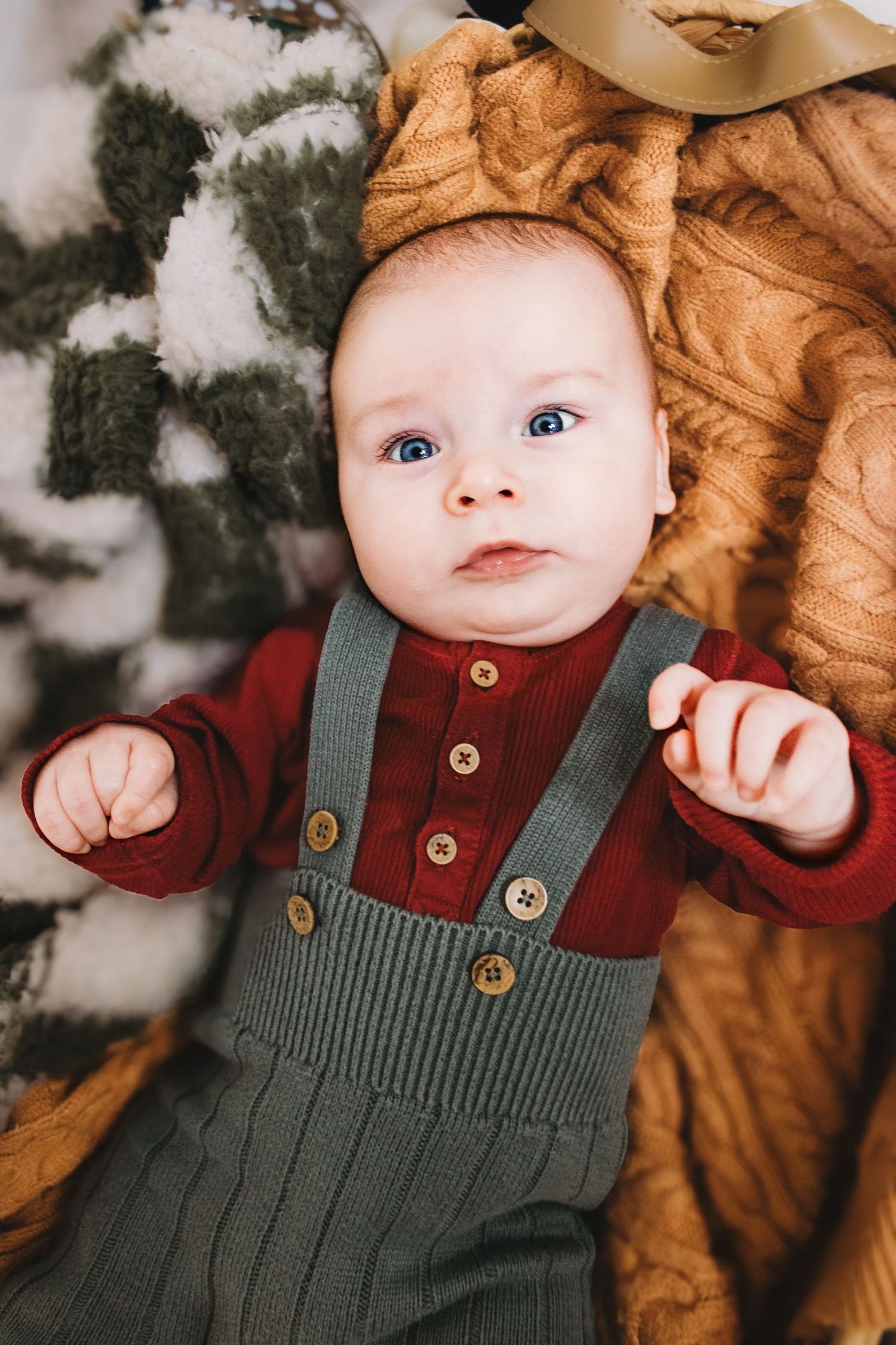 Baby wearing green knit overalls and burgundy shirt lies on soft mustard yellow blanket in winter setting.