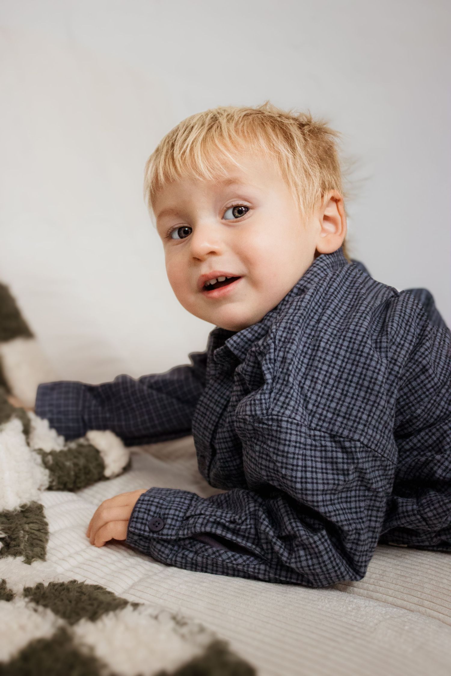 A toddler in a navy checkered shirt lies on a light-colored surface playing with gray material.