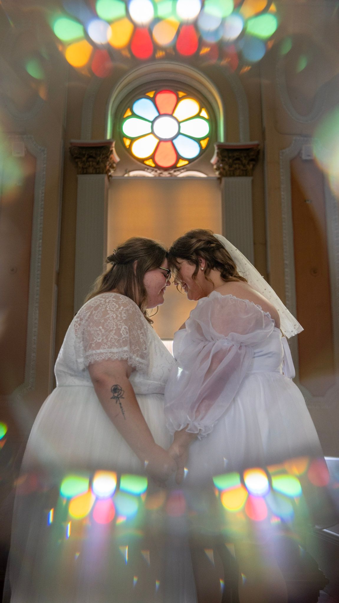 Wedding couple embracing under stained glass windows casting colorful reflections in an elegant venue.