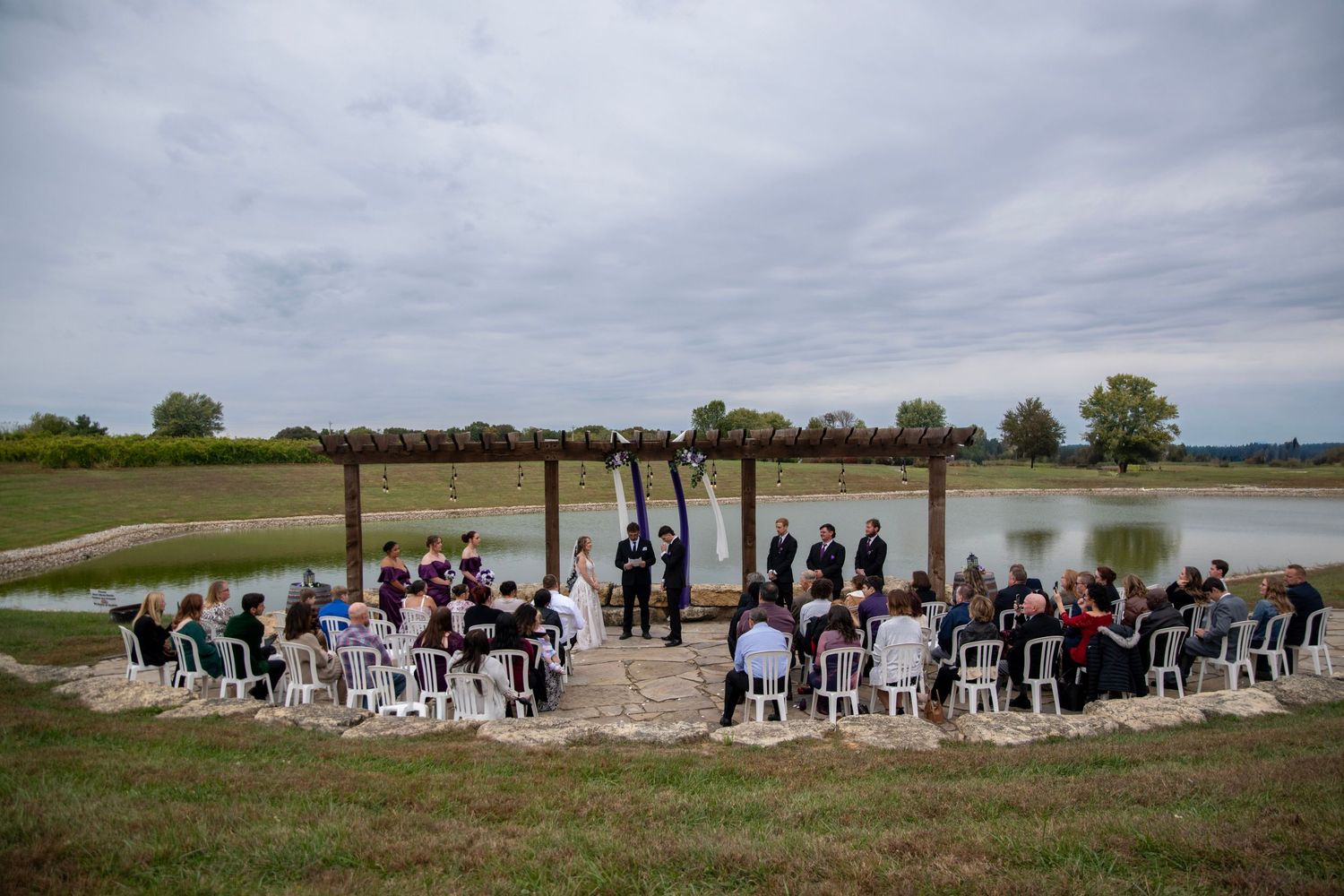 Wide angle view of lakeside wedding ceremony setup with guests seated under pergola.