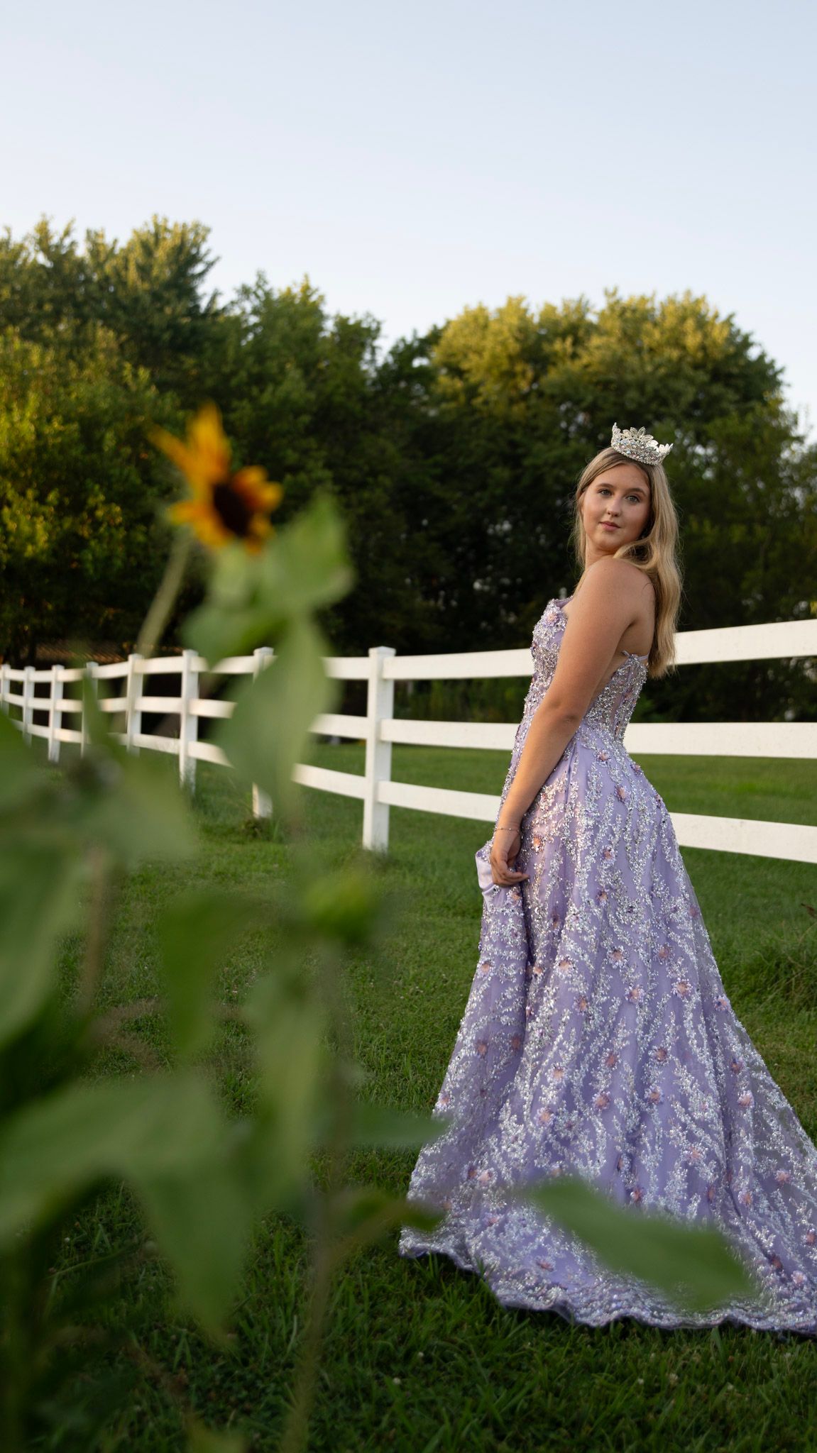 Lavender ball gown with sparkles photographed by a white fence with sunflowers in view.