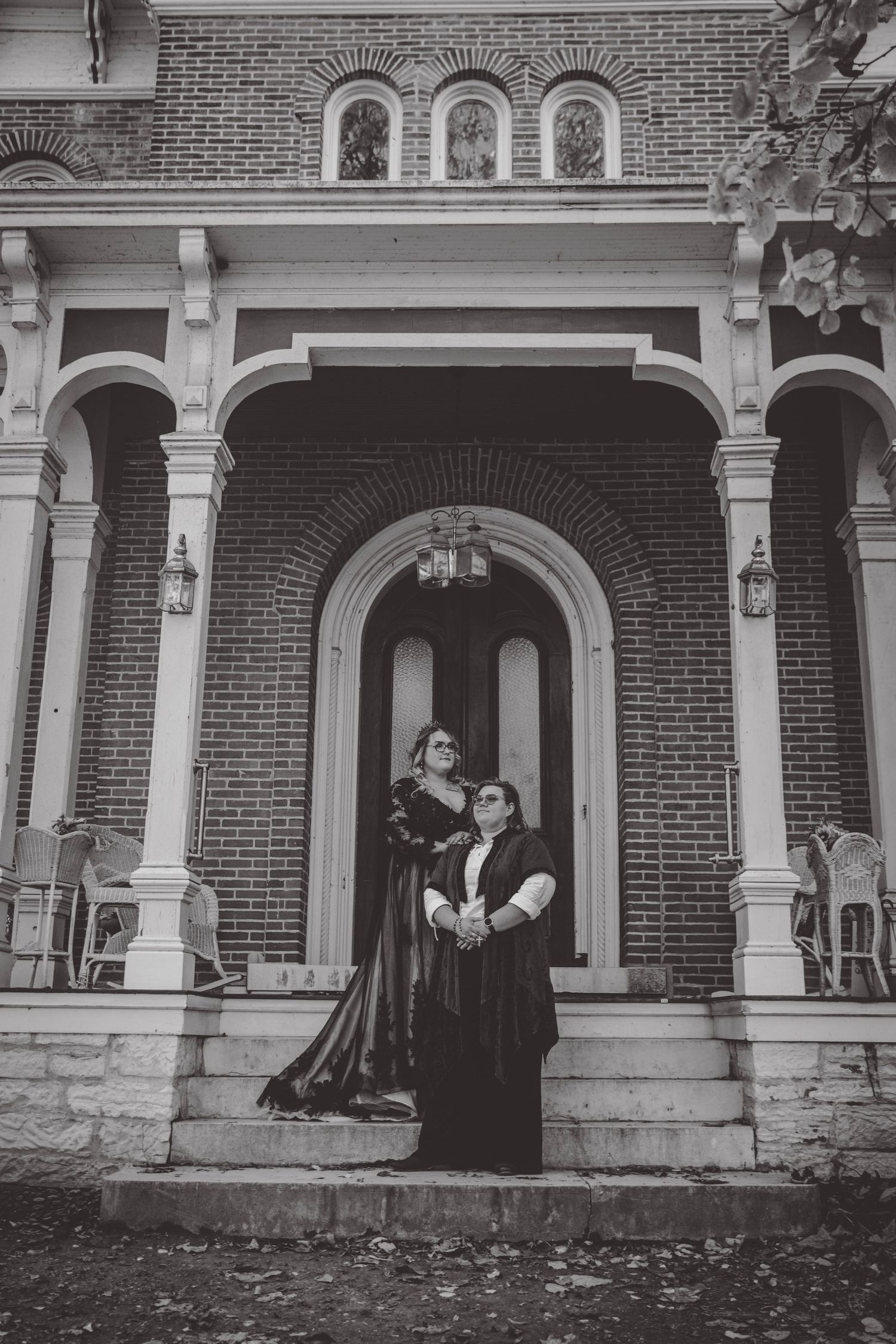Gothic couple poses on steps of historic brick building with arched entrance.