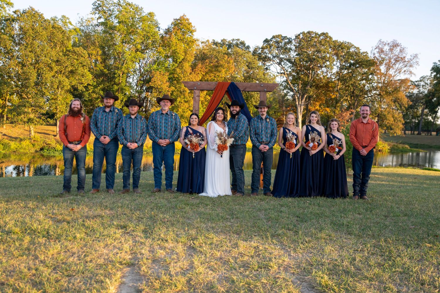 Wedding party stands in line against autumn trees during golden hour photography session.