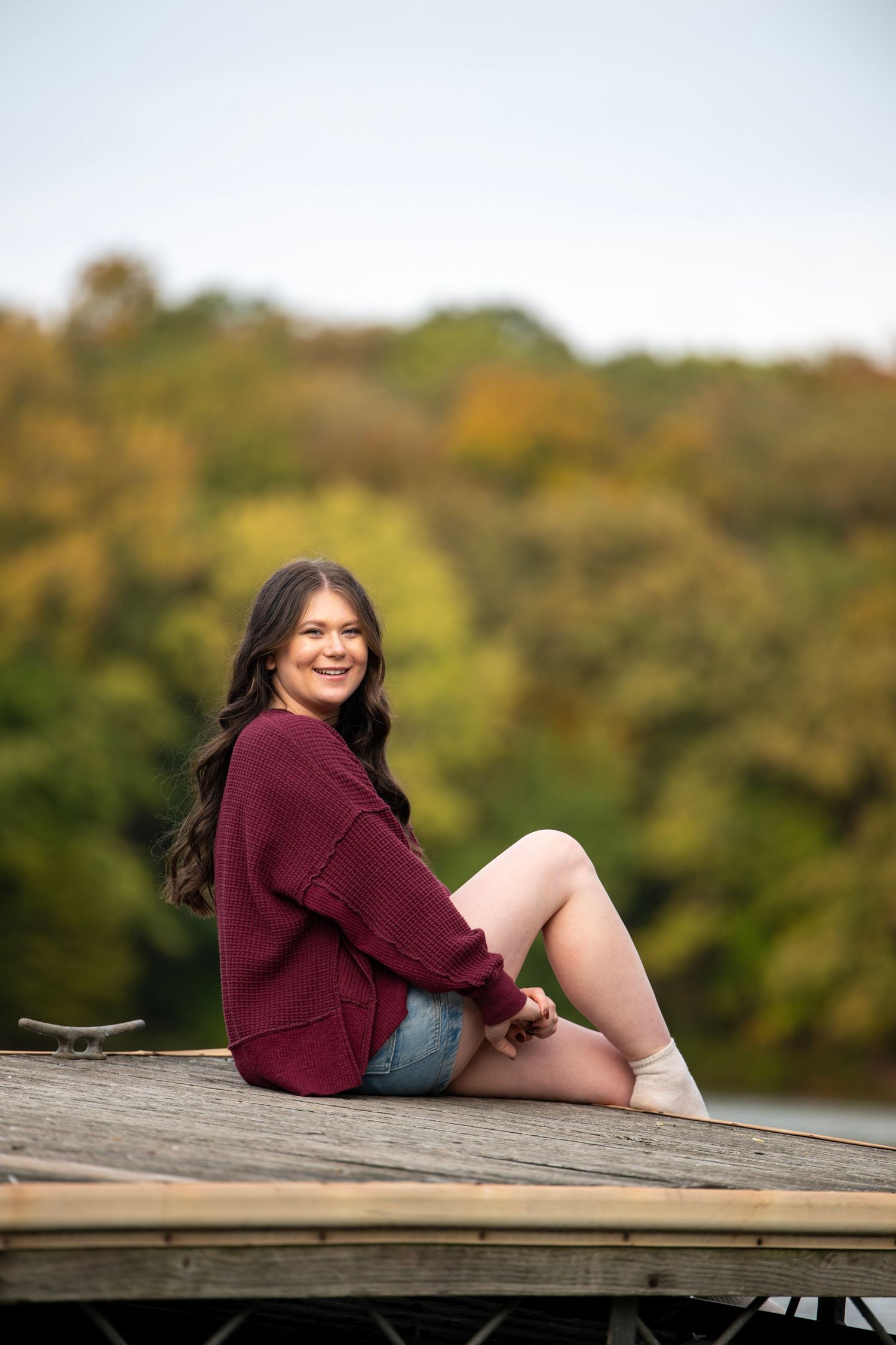 Person in burgundy sweater sits on wooden dock with fall scenery in background.