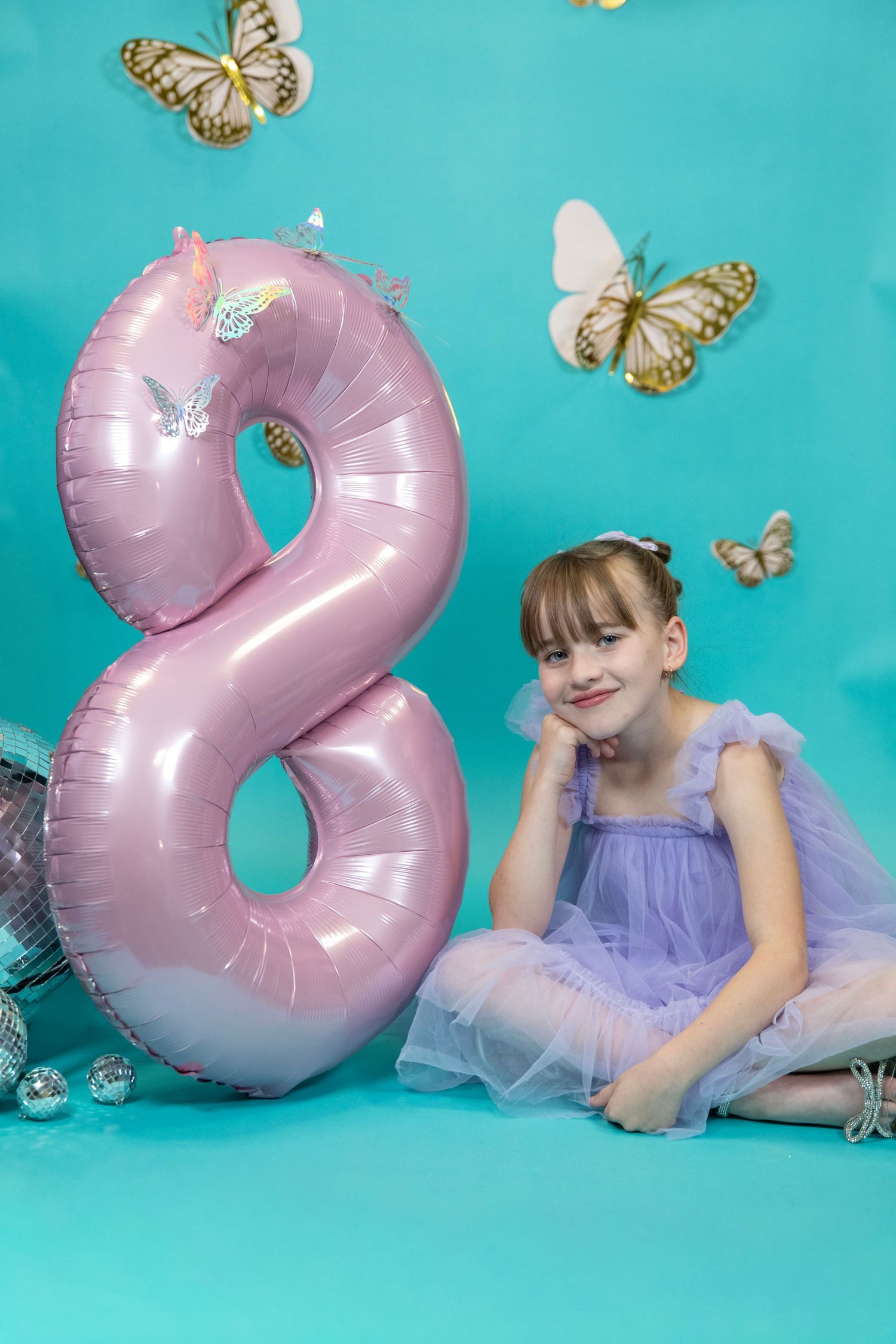 Playful pose with pink number 2 balloon and butterfly decorations on turquoise background.