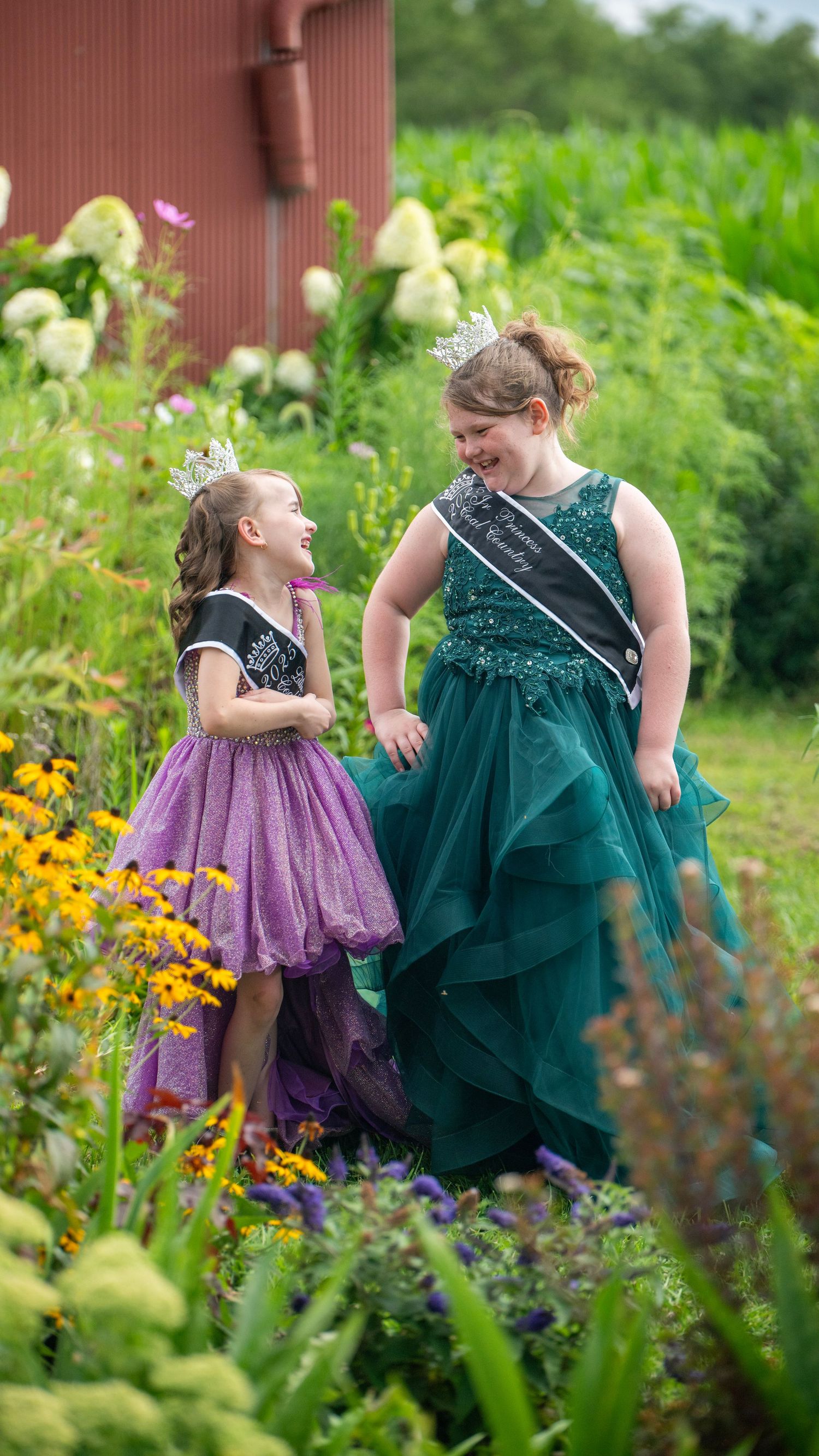 Two people in formal dresses share a joyful moment in a garden filled with colorful flowers and greenery.