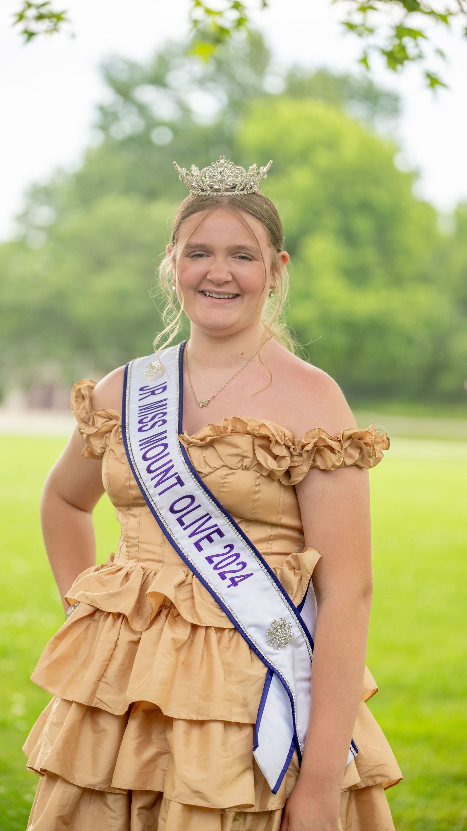 Pageant contestant in a gold ruffled dress with sash and tiara posing outdoors.