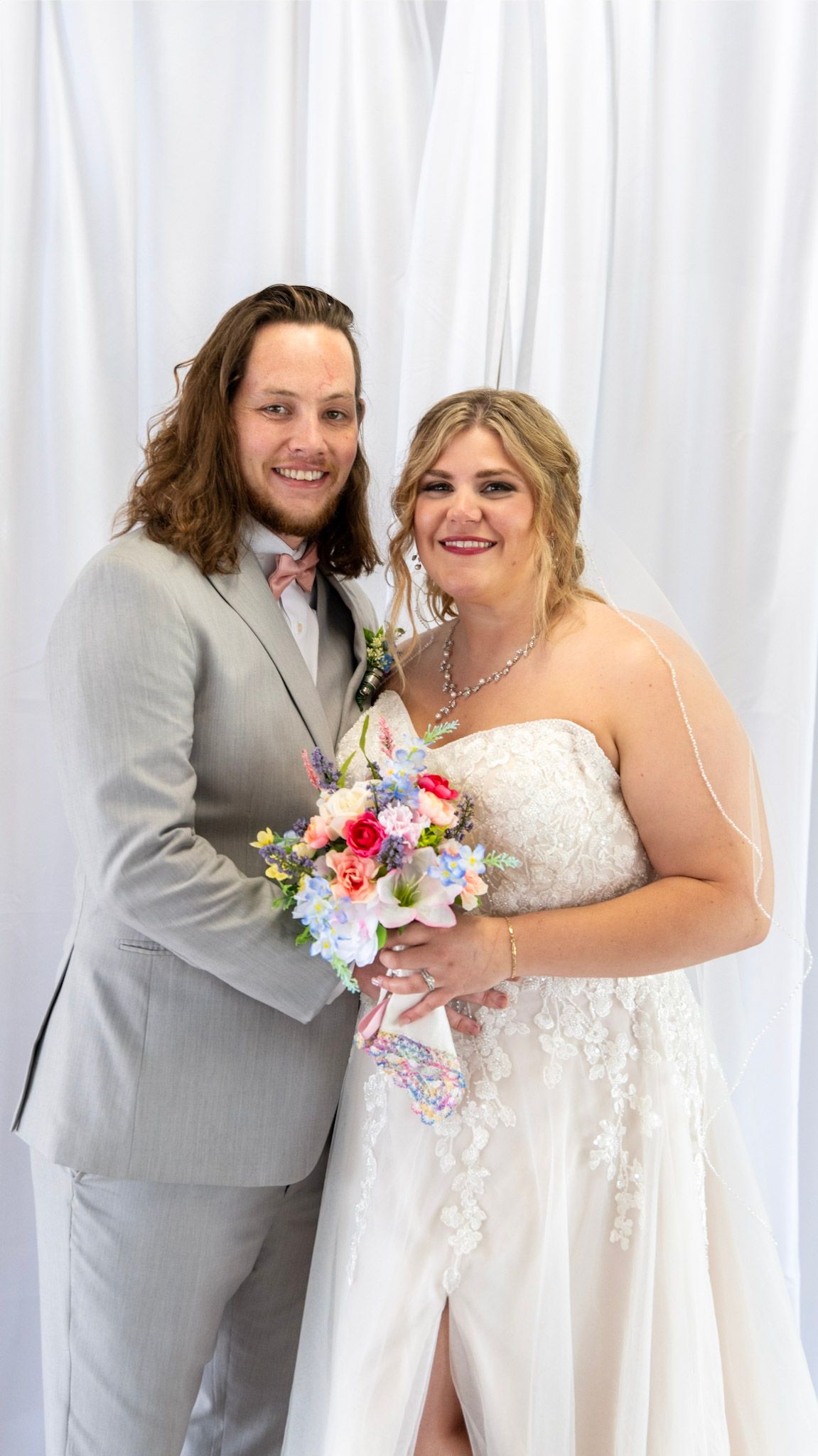 Wedding couple poses together against white backdrop with colorful floral bouquet.