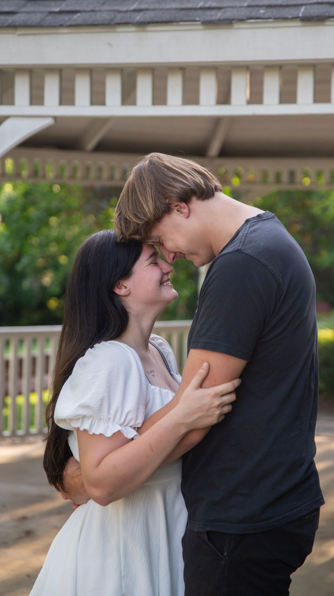 A romantic couple embraces in front of a white gazebo on a sunny day.
