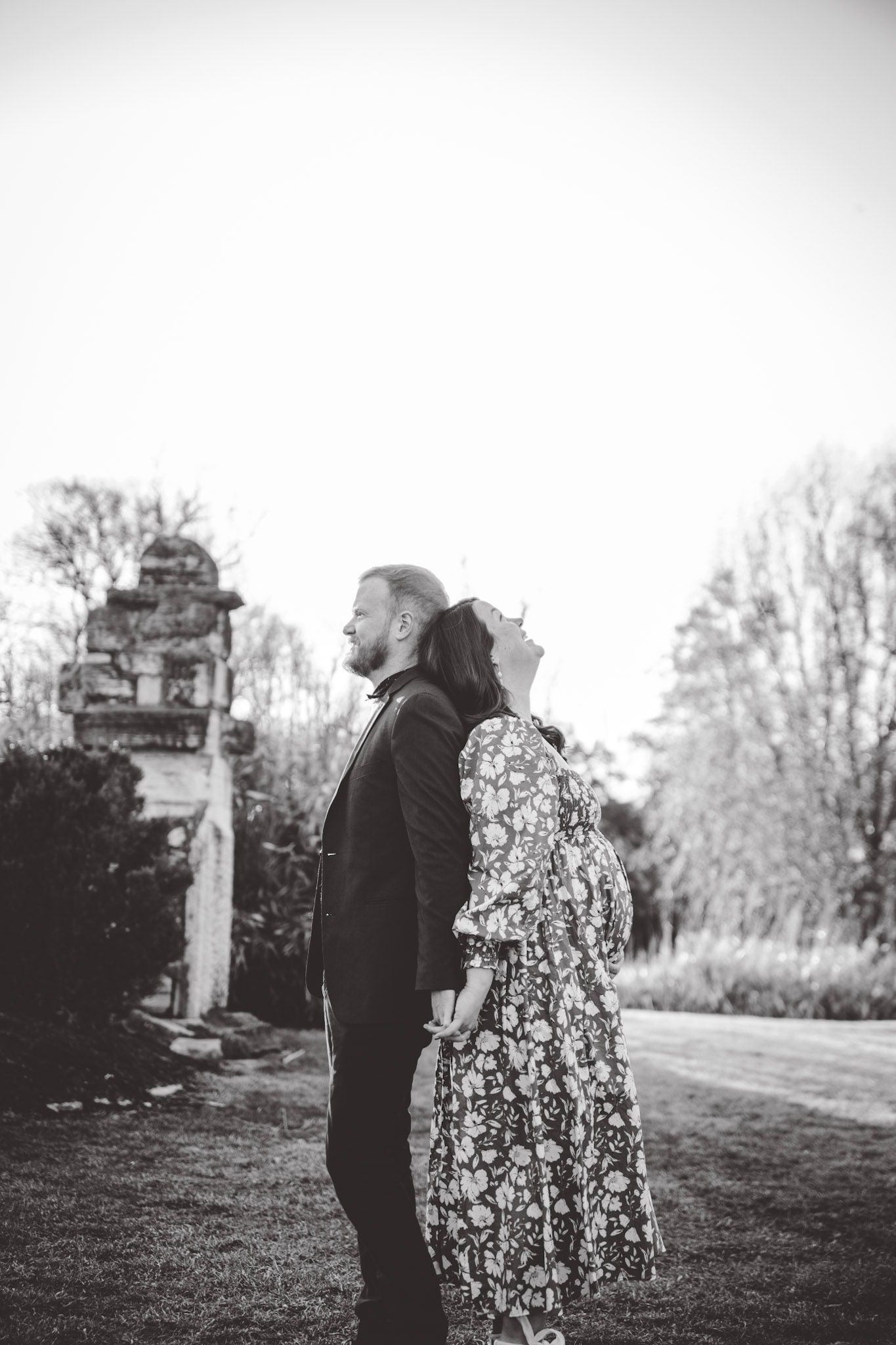 Black and white portrait of couple in romantic pose against rustic stone wall and trees.