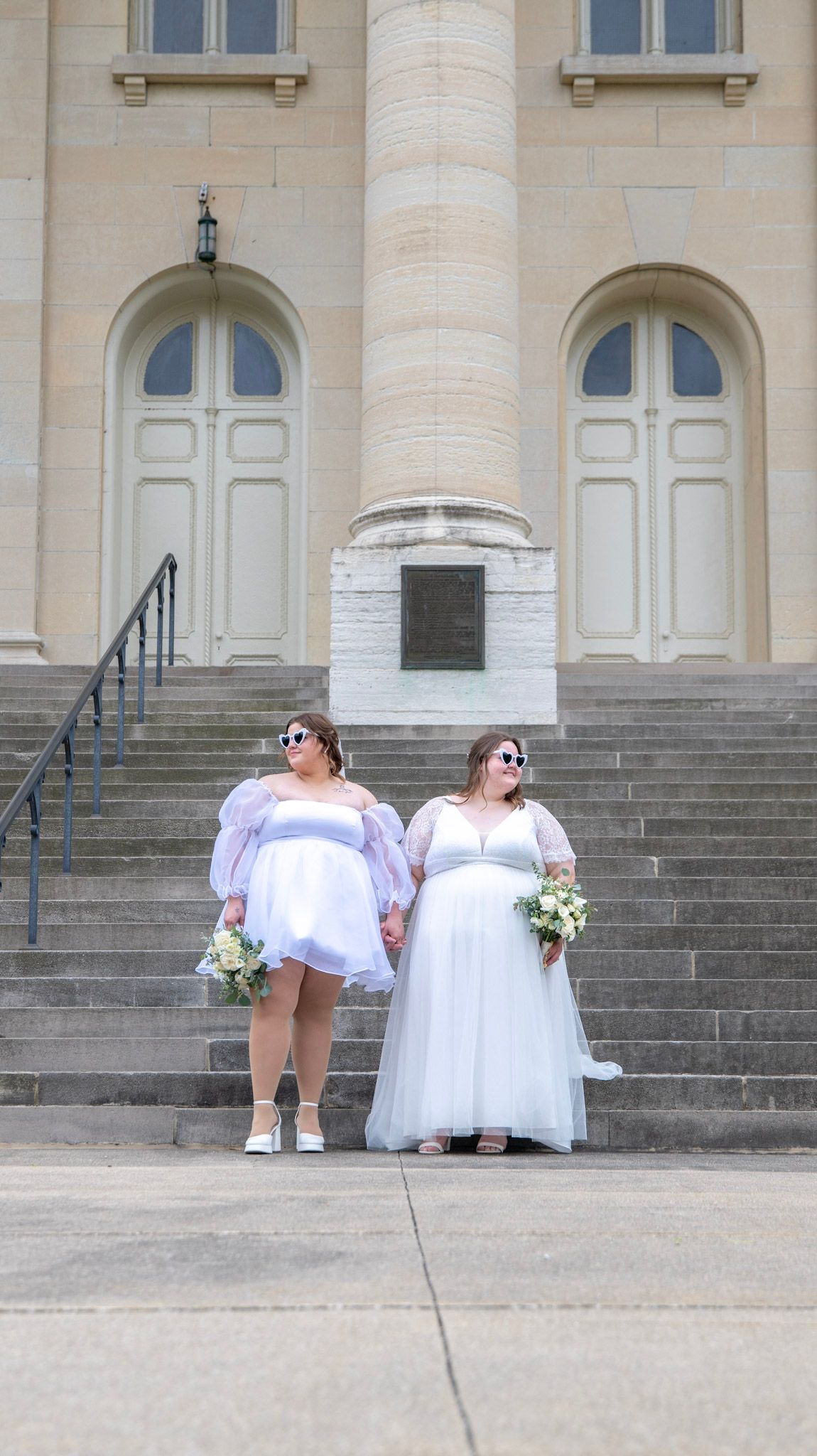 Two people in flowing white dresses pose playfully on wide stone steps of a classical building.