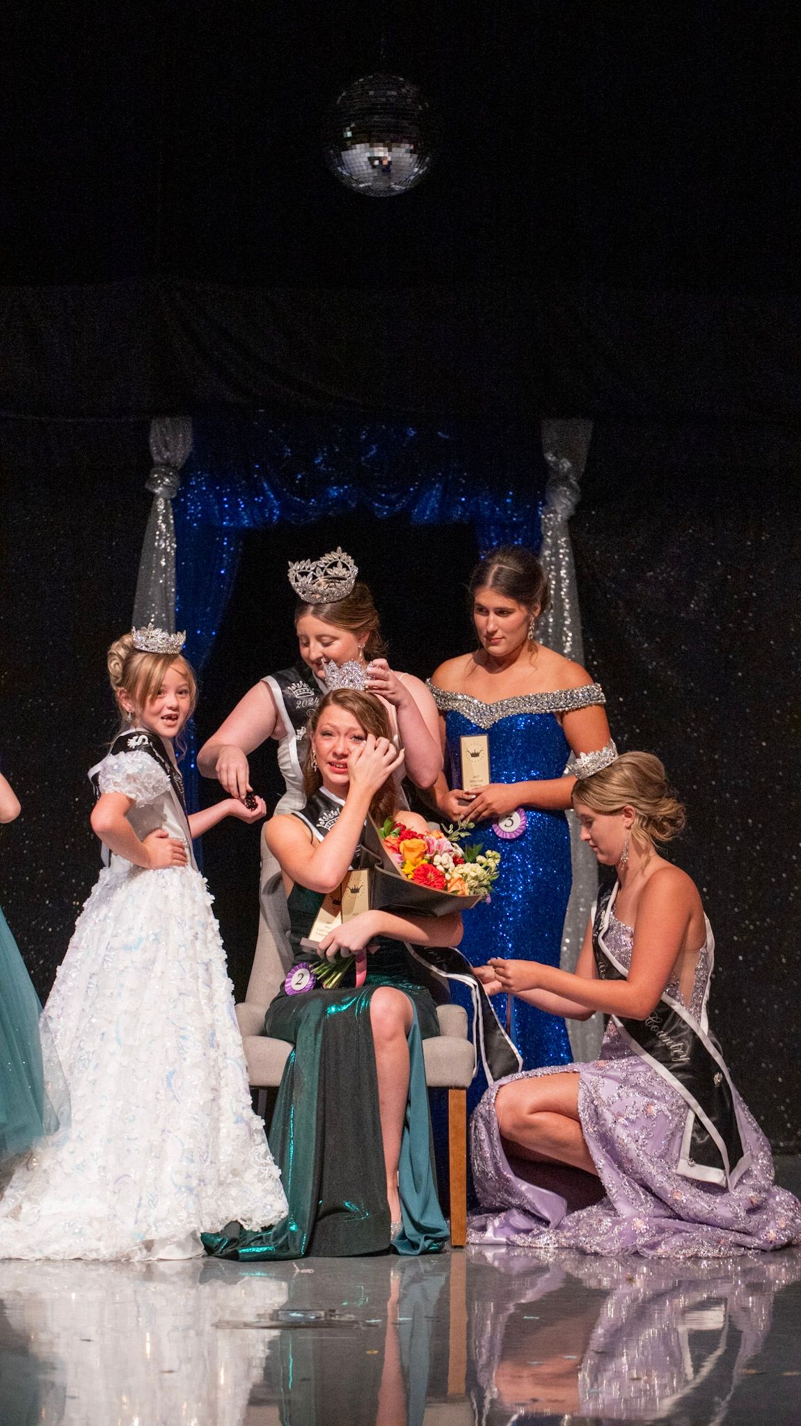 Emotional pageant participants share a group moment on stage in formal gowns.