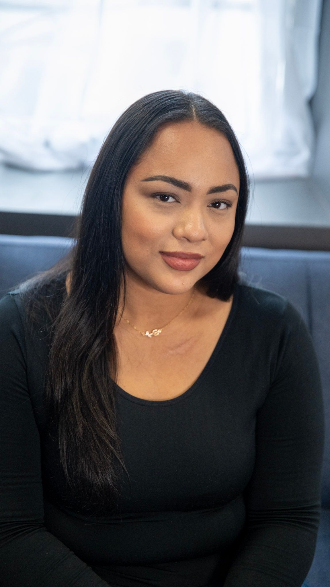 Portrait of a woman in black top sitting on grey couch near window with natural lighting.