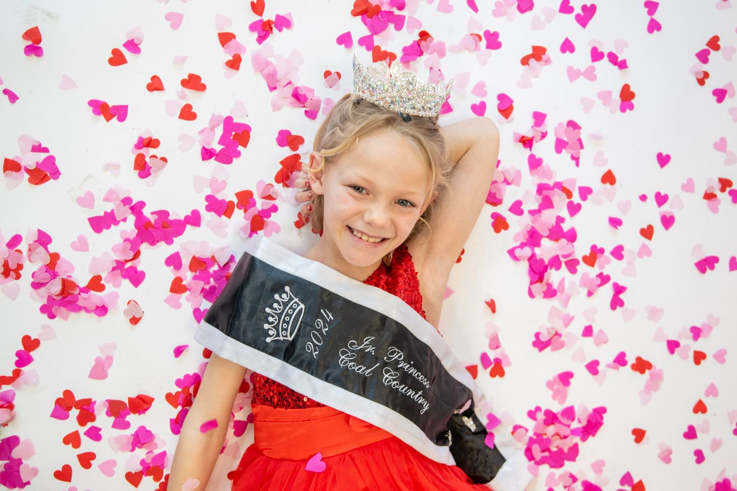 Pink confetti falls over festive scene with red dresses and sparkly crowns.