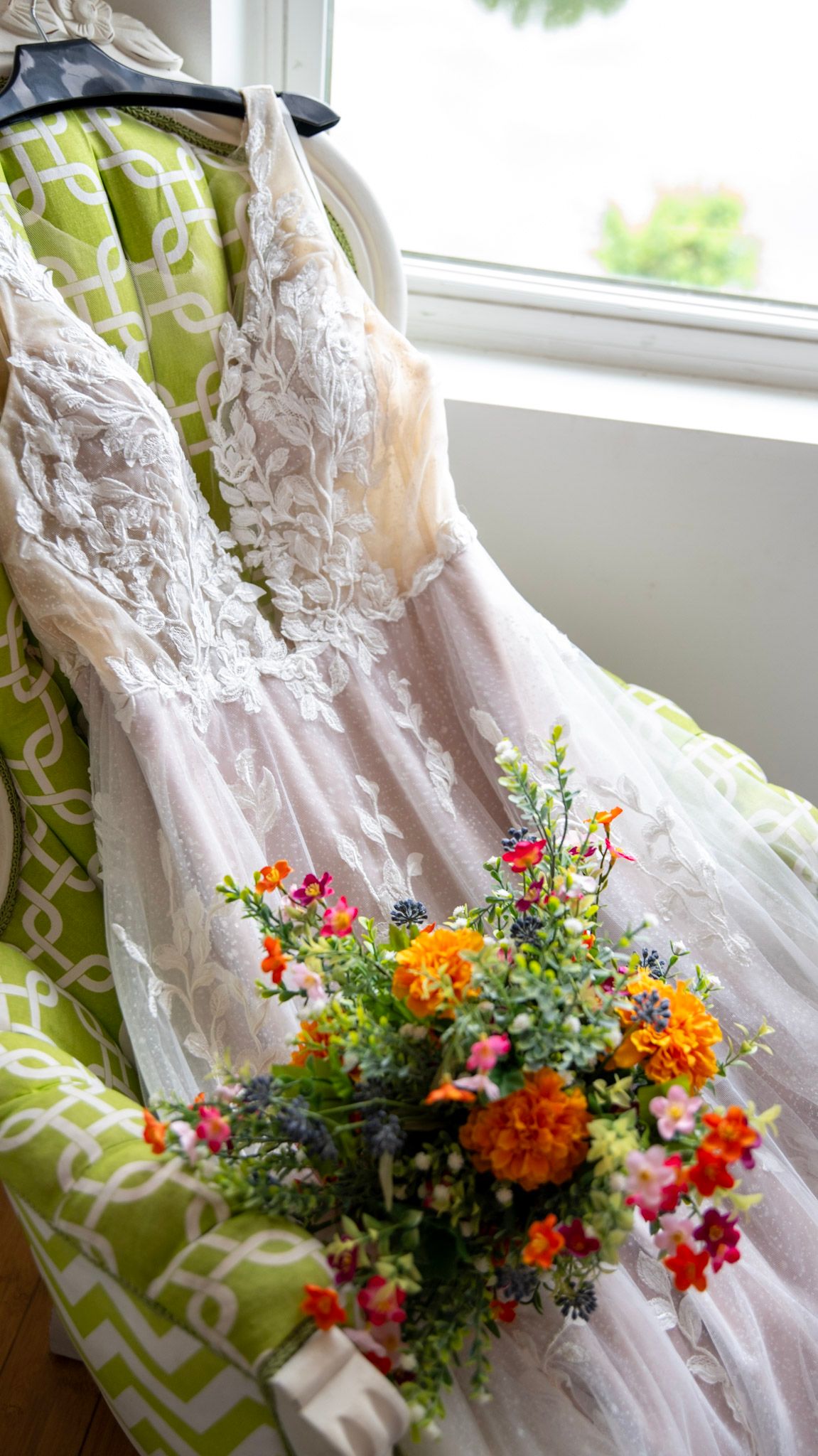 White lace wedding dress hangs by window with vibrant orange and red floral bouquet.