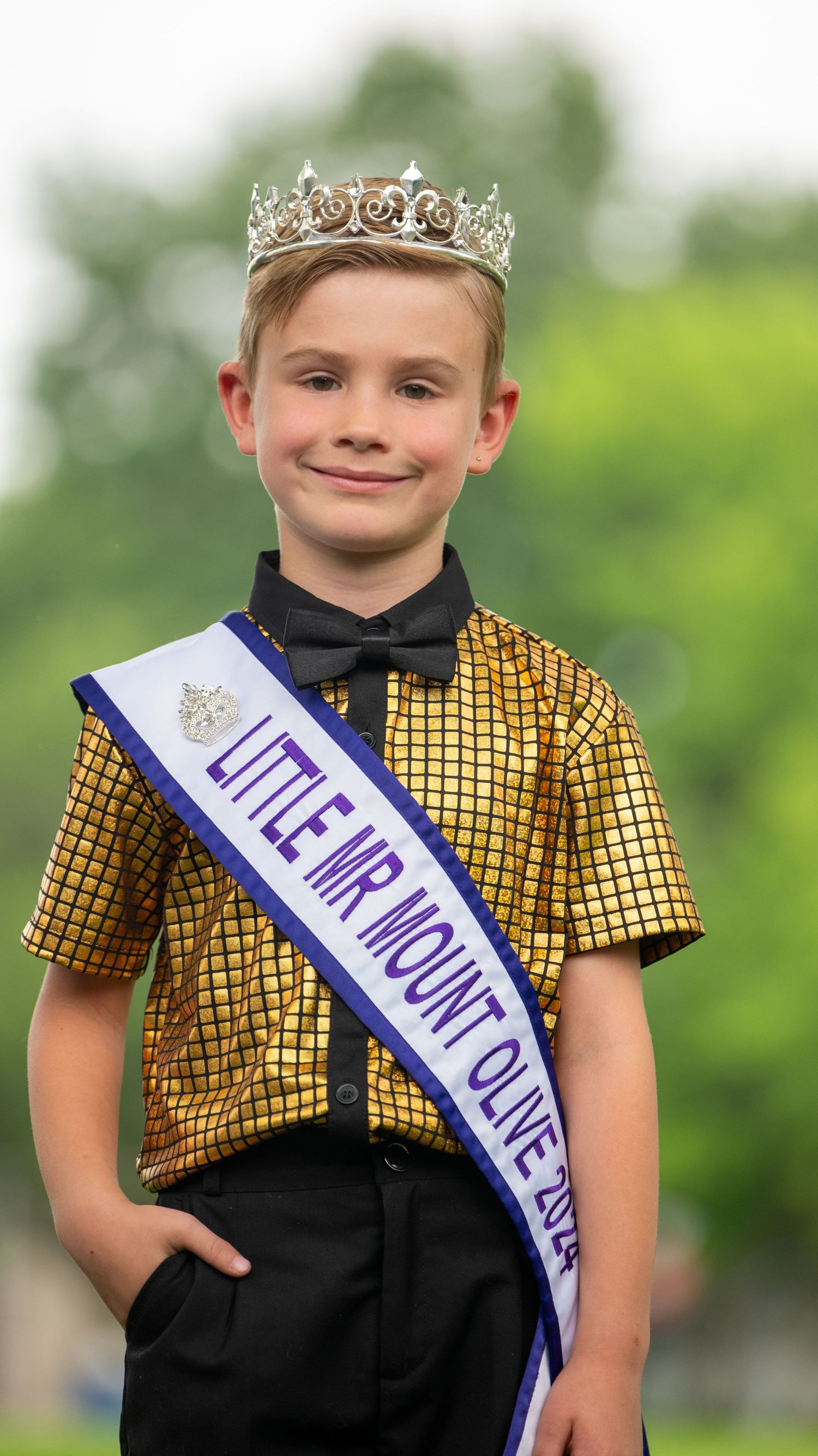 Young pageant contestant in gold and black outfit wearing crown and sash poses outdoors.