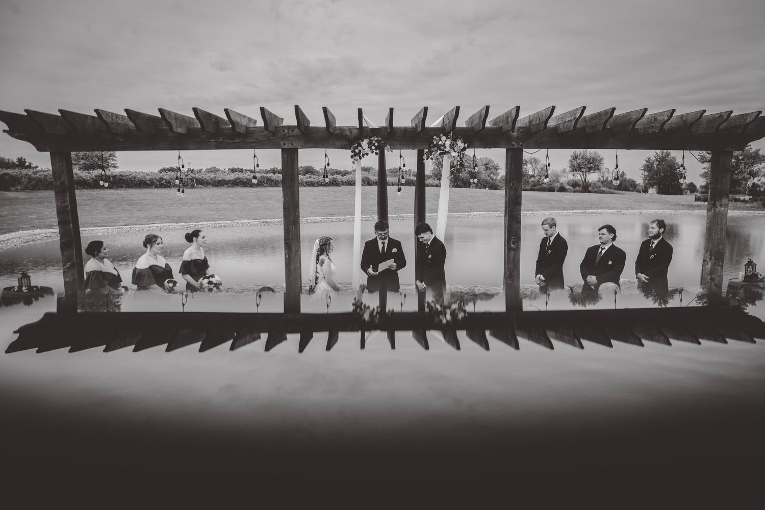 Black and white artistic photo of wedding party framed by wooden pergola structure.