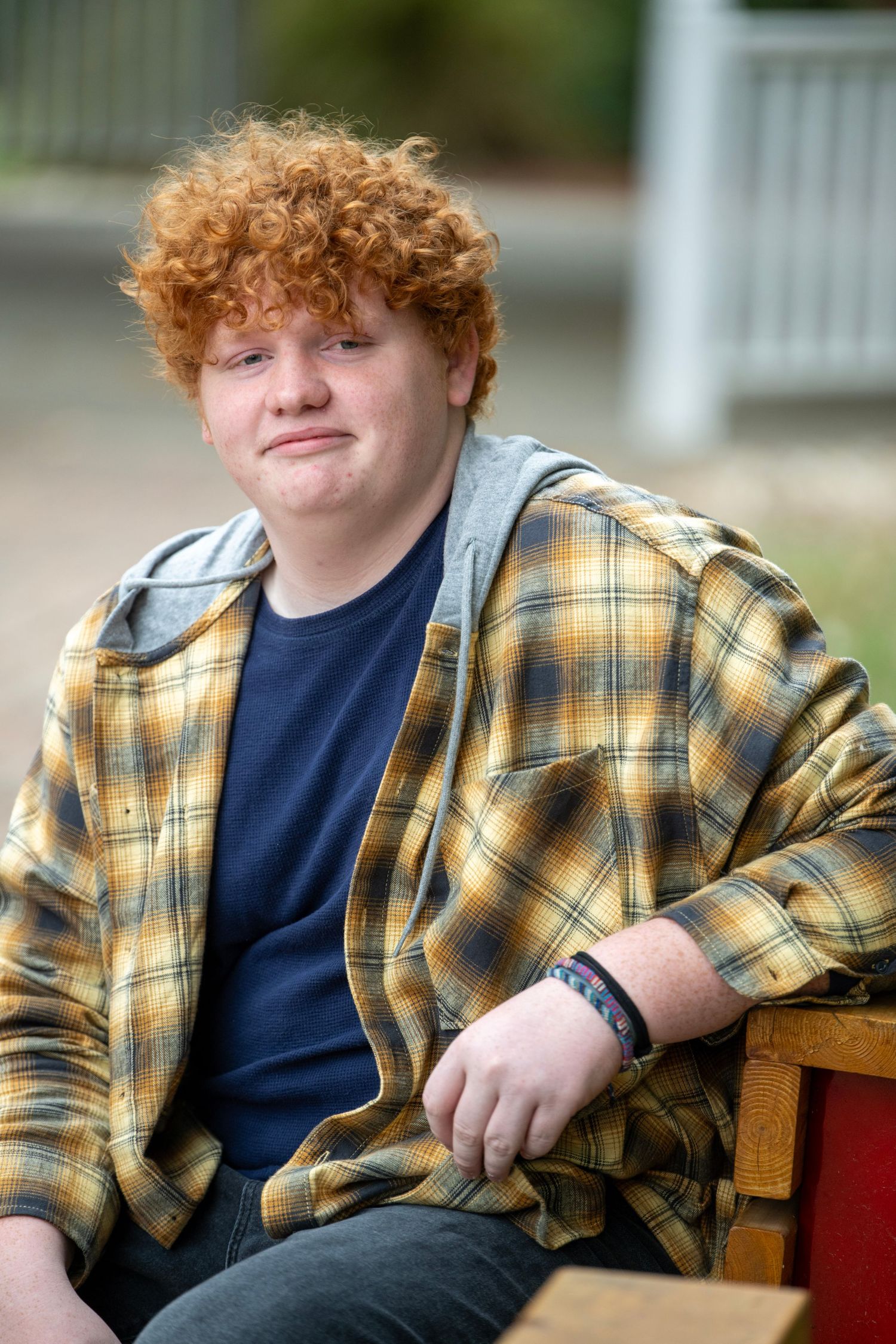 Relaxed portrait on wooden bench wearing plaid jacket and navy shirt.