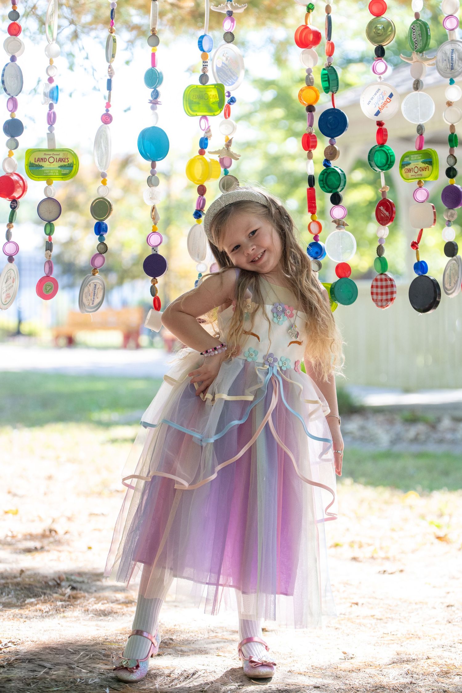 Child in fairy costume poses beneath colorful hanging decorations in an outdoor setting.