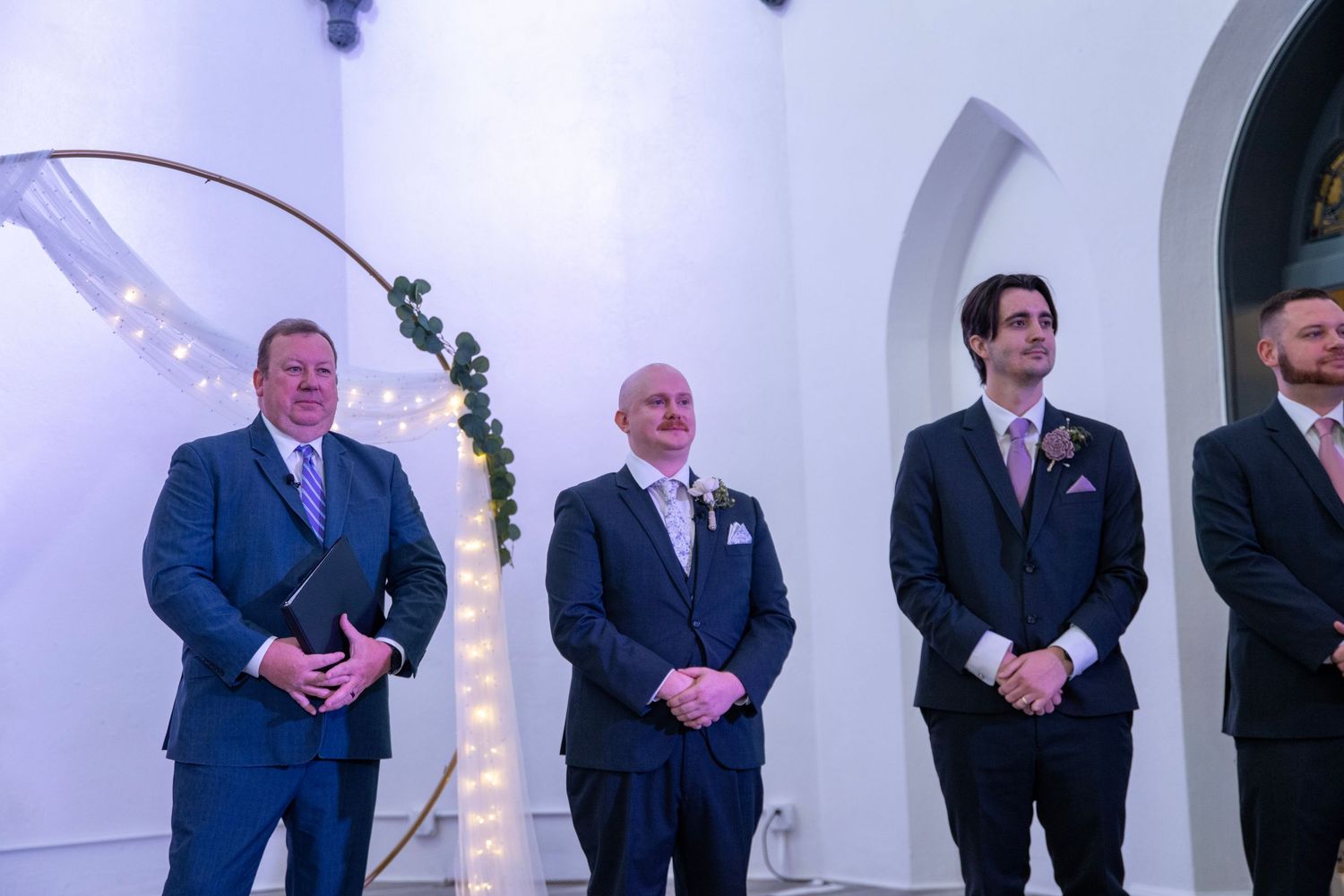 Groomsmen in navy suits stand in line during wedding ceremony.