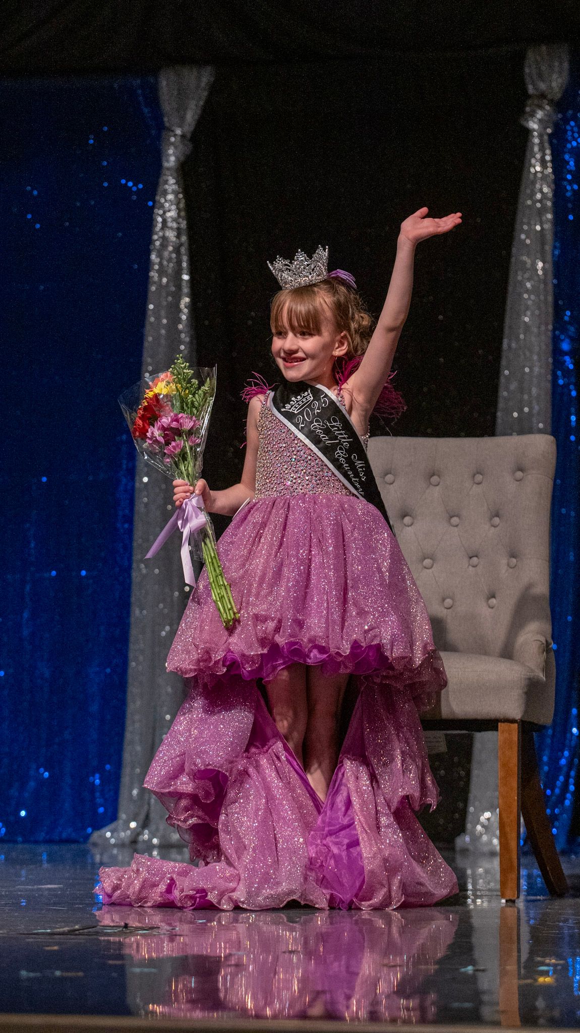 Young pageant contestant in sparkly pink high-low dress and crown waves while holding bouquet on stage.