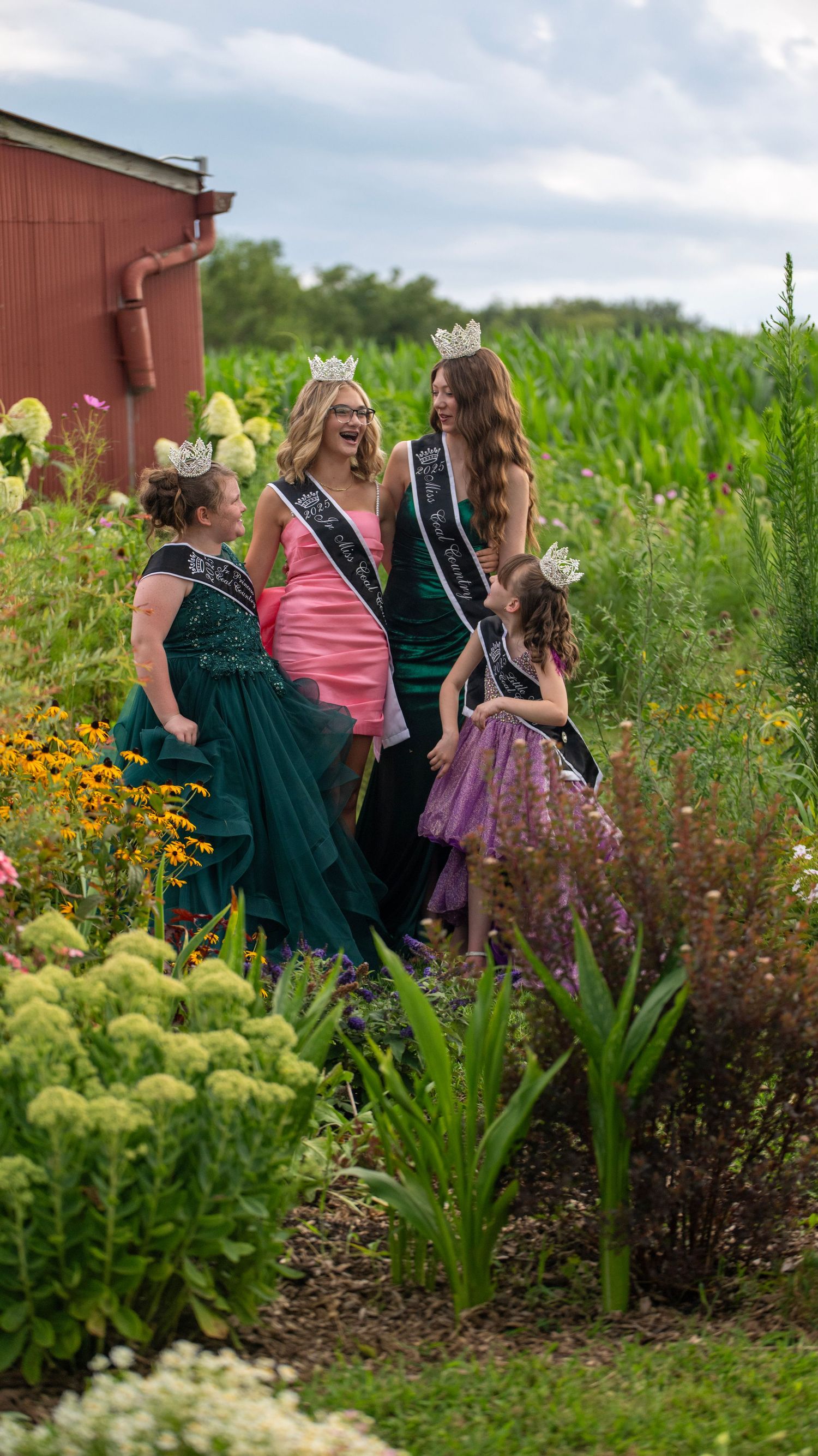 A group poses together in elegant dresses among blooming flowers and greenery near a red barn.