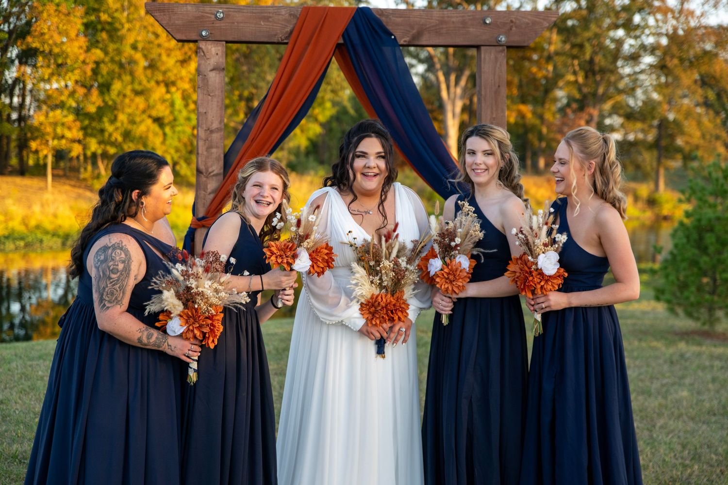 Wedding party in navy dresses holds autumn-colored bouquets against wooden arch backdrop.
