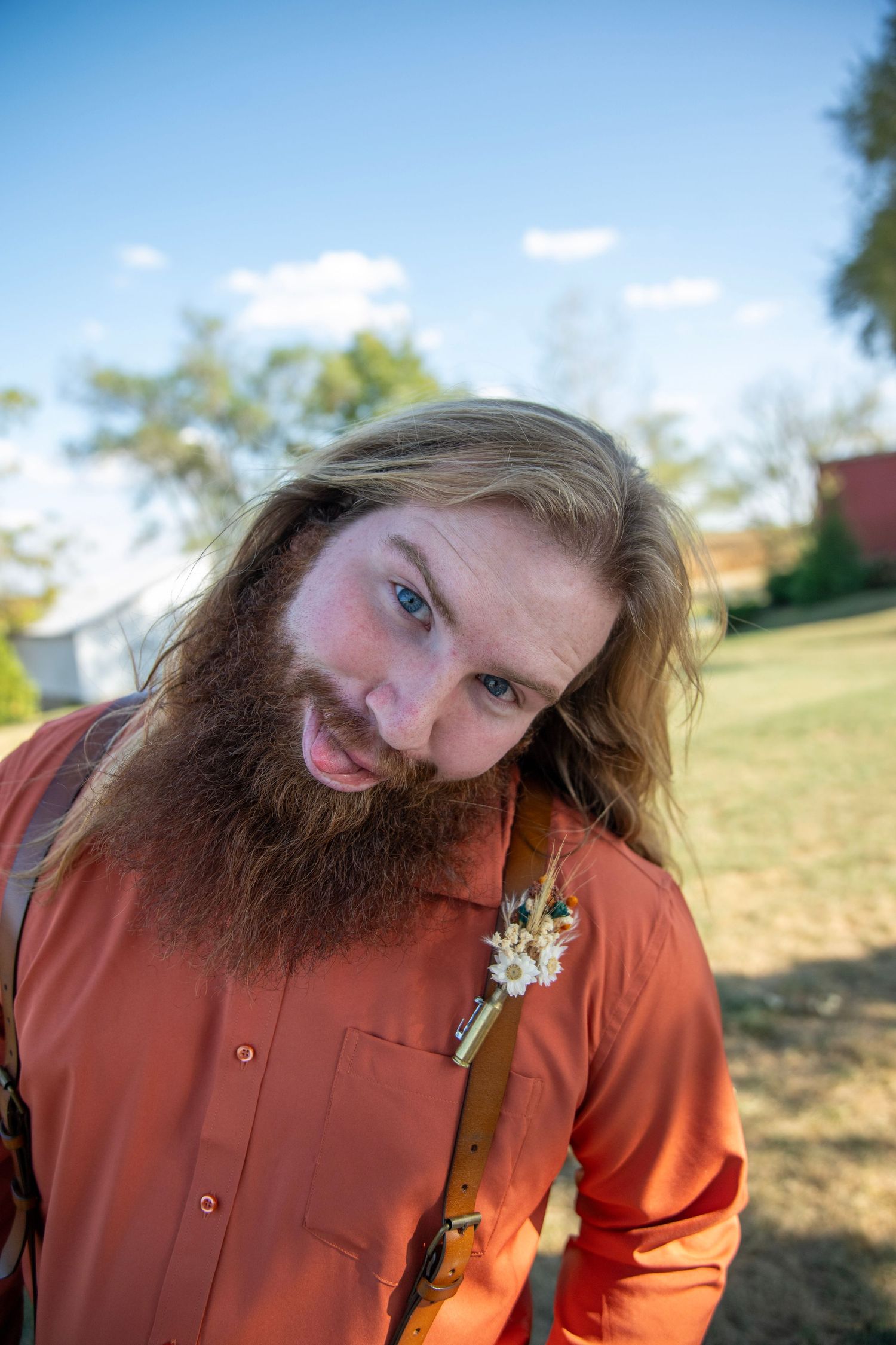 Person in orange shirt with long beard and floral accessory poses outdoors.