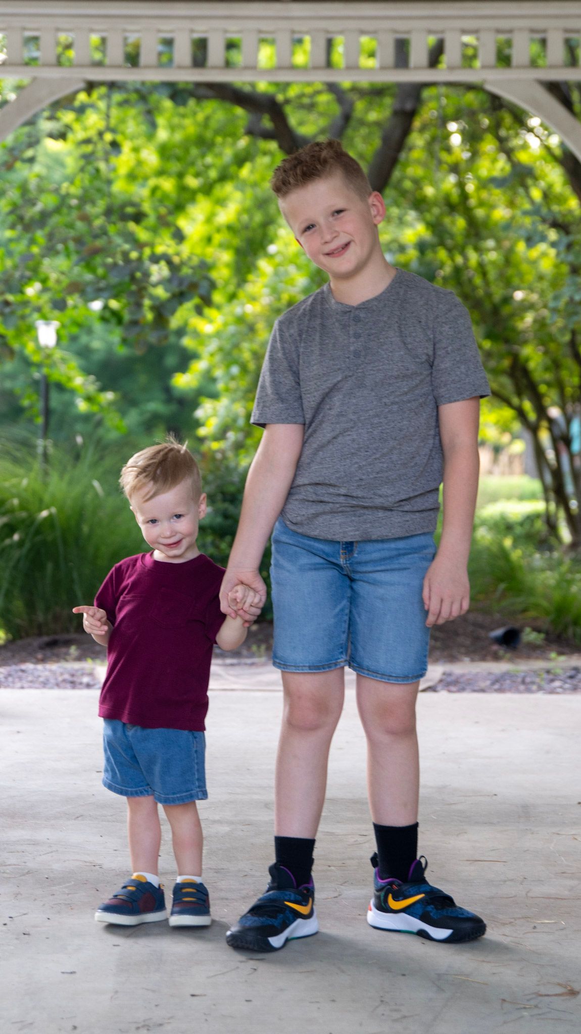 Two people in casual attire standing together on a paved walkway surrounded by greenery.
