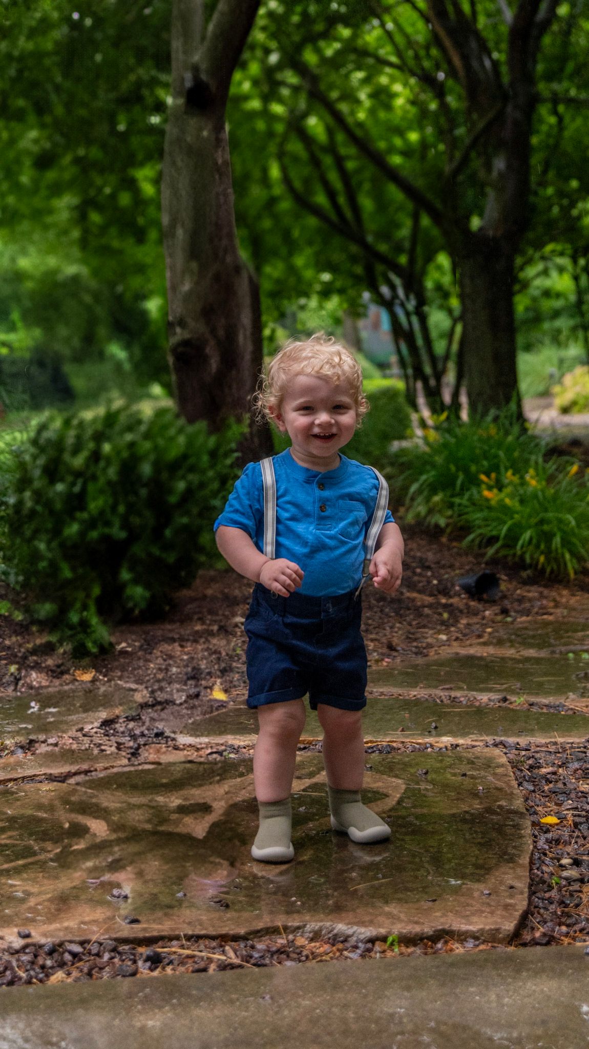 Small child in blue clothing standing on a stone surface in a lush garden setting.