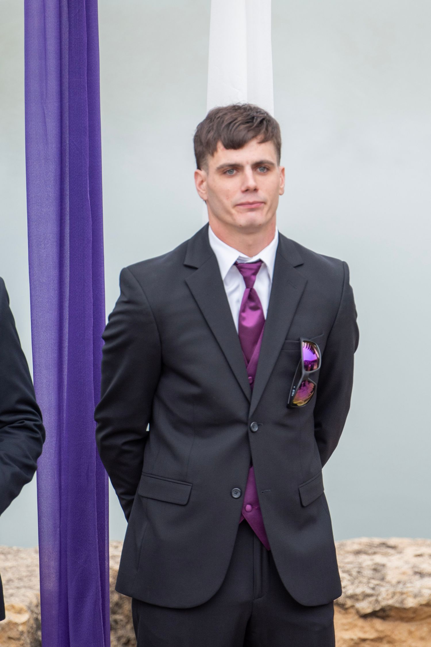 Person in black suit with purple tie standing between decorative purple pillars.