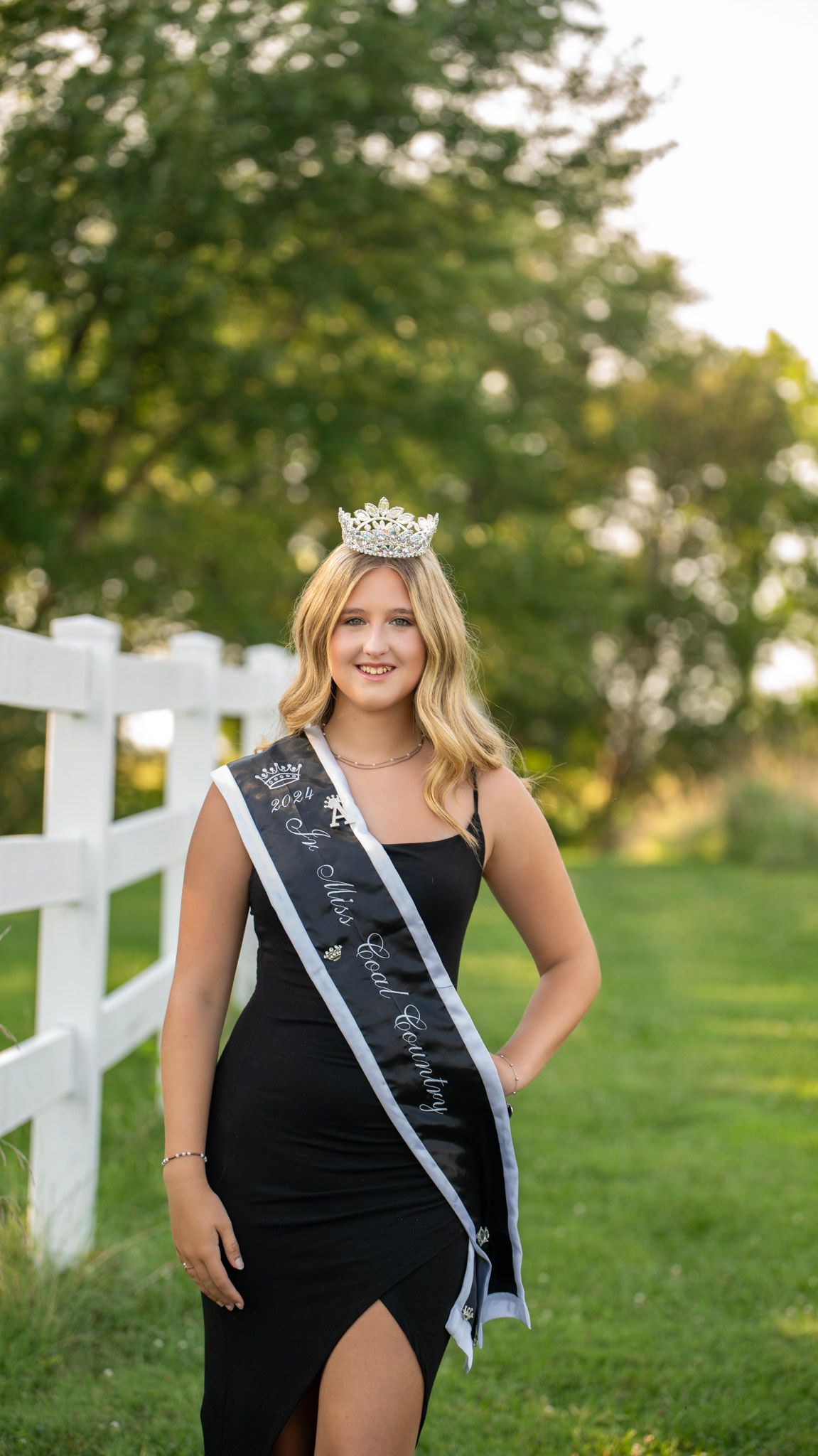 A pageant winner in a black dress poses by a white fence wearing a crown and sash.
