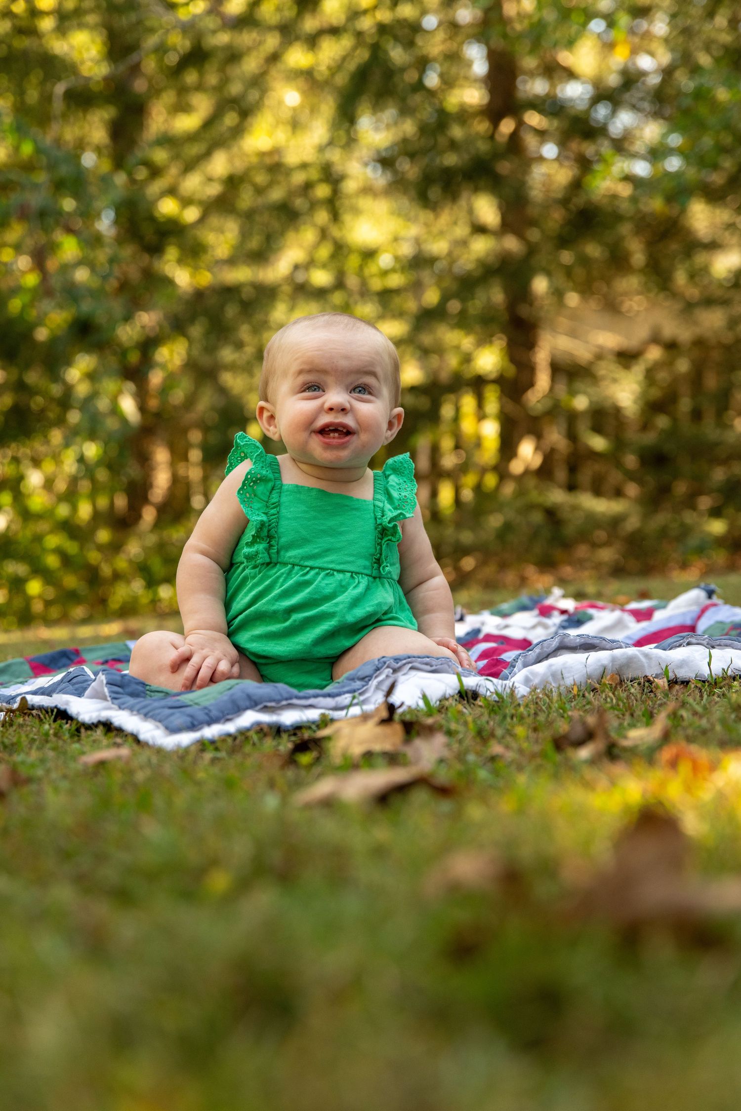 Happy baby in bright green romper sitting on colorful blanket outdoors.