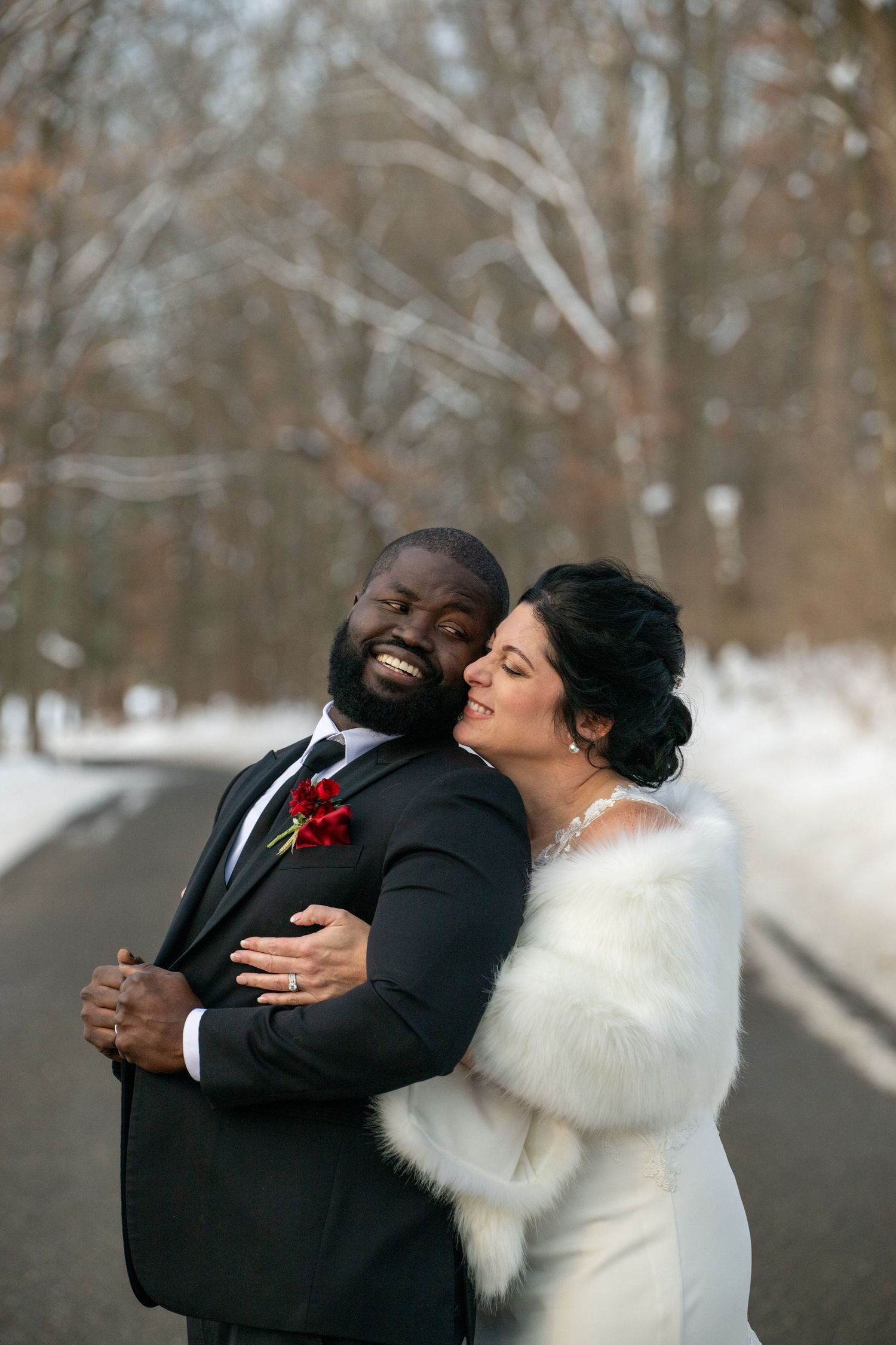 Winter wedding couple embracing on snowy path with bare trees in background.