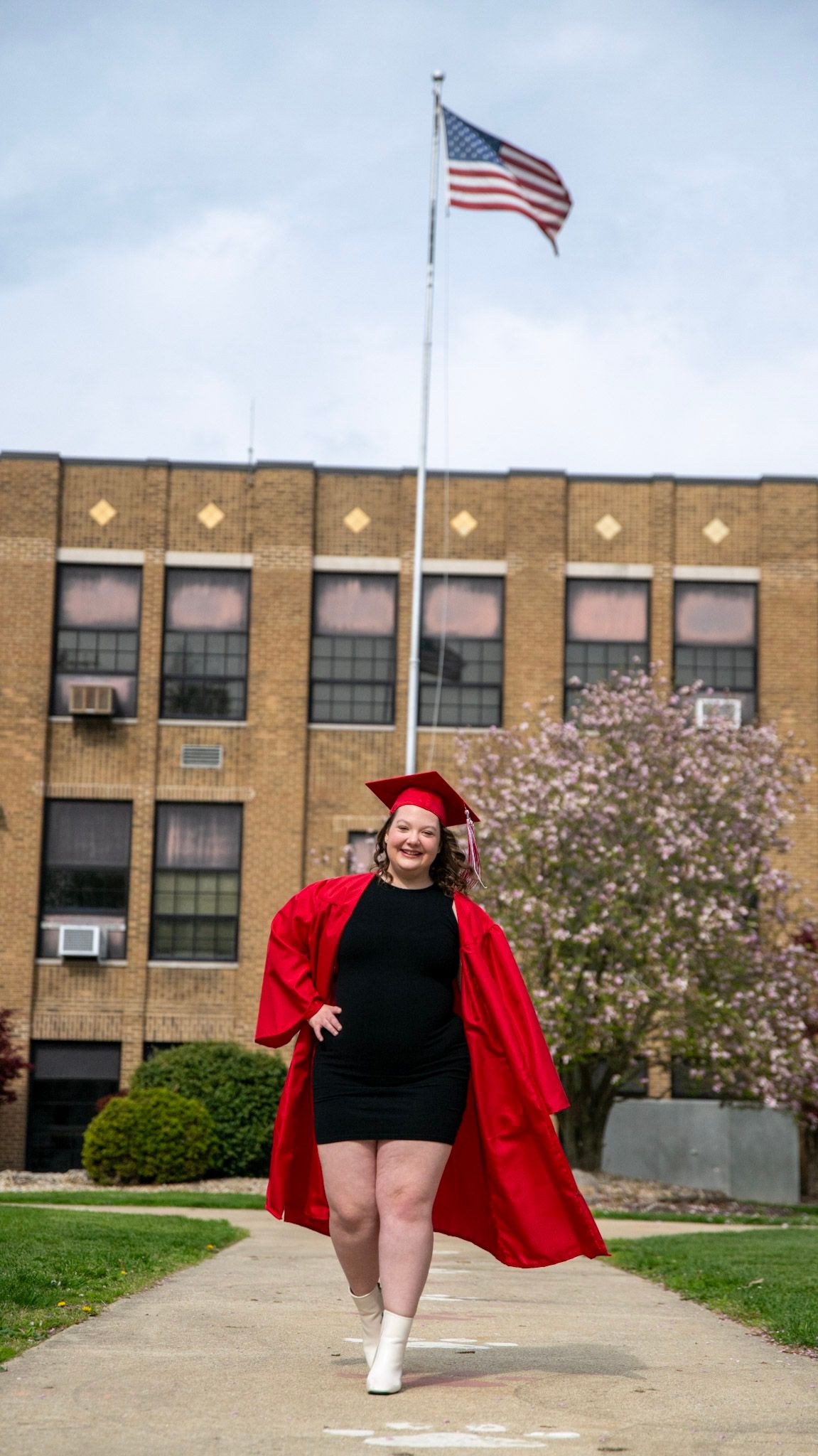 Graduate in red cap and gown poses playfully in front of brick school building with American flag.