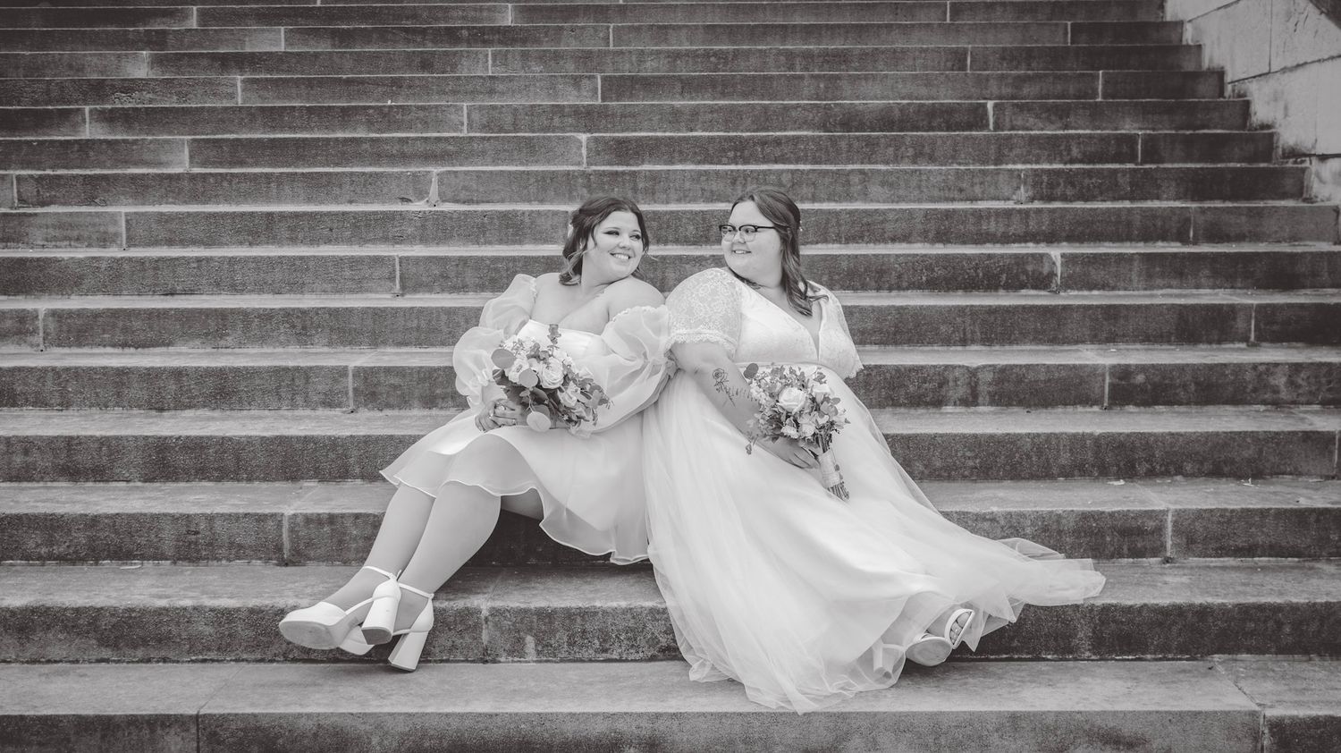 Wedding couple in white dresses posing playfully on stone steps in black and white.
