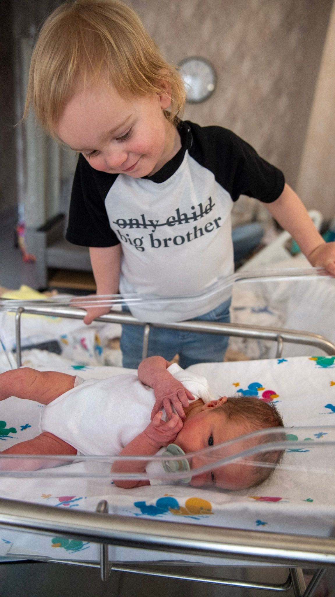 Child wearing a big brother shirt looking down at a hospital bassinet.