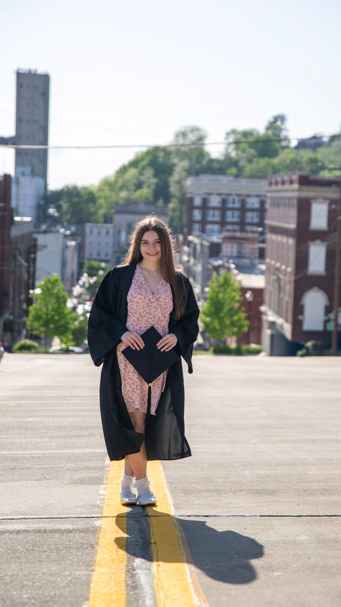 Graduate in black gown and floral dress poses on yellow parking lines with cityscape behind.
