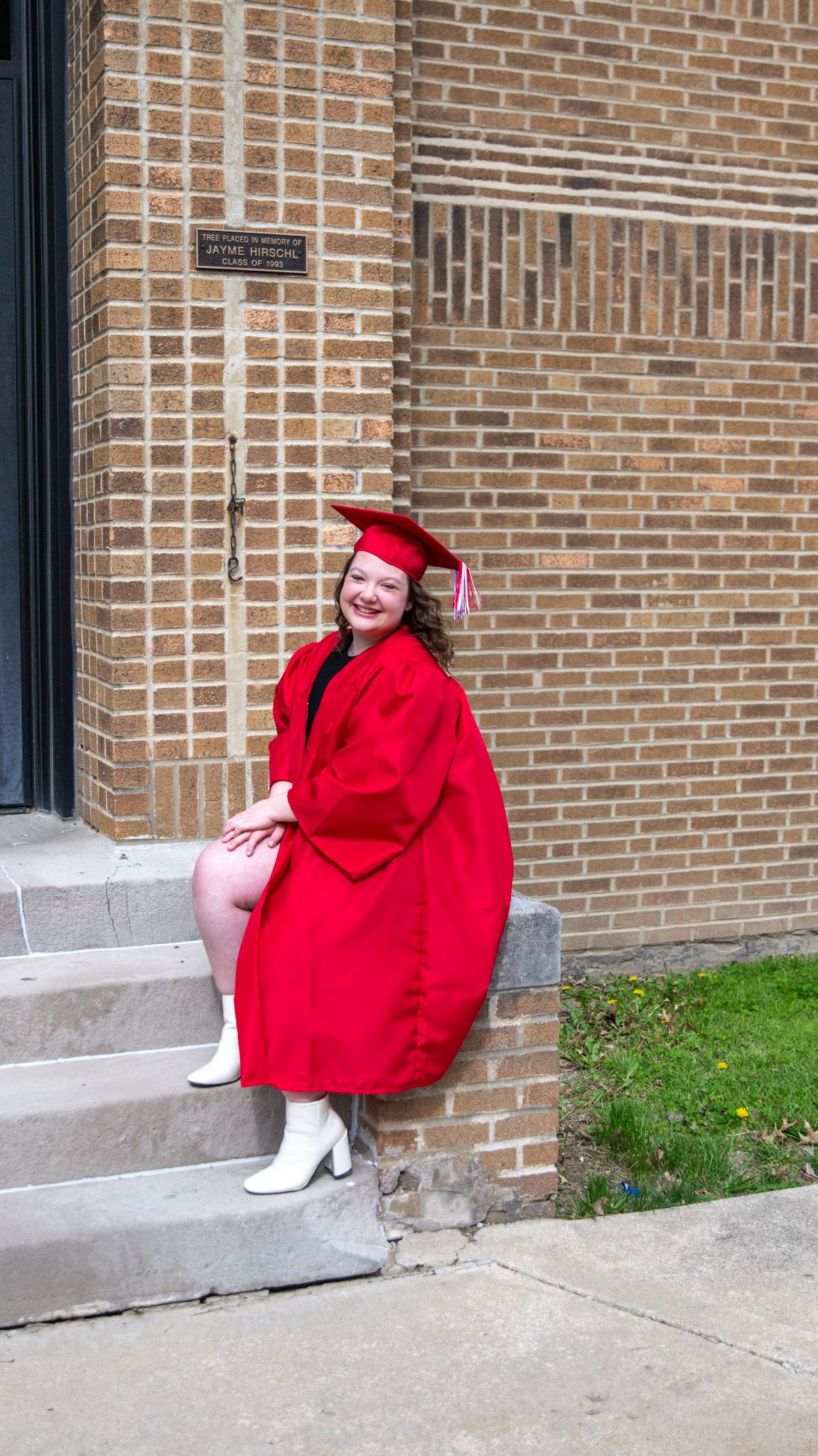 Graduate in bright red cap and gown sits on concrete steps by brick wall.