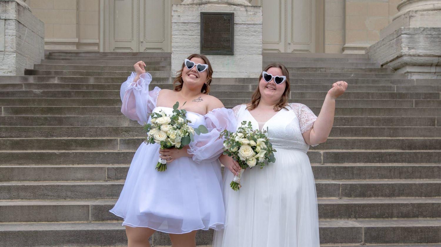 Wedding party in white dresses poses playfully on grand stone steps of historic building.