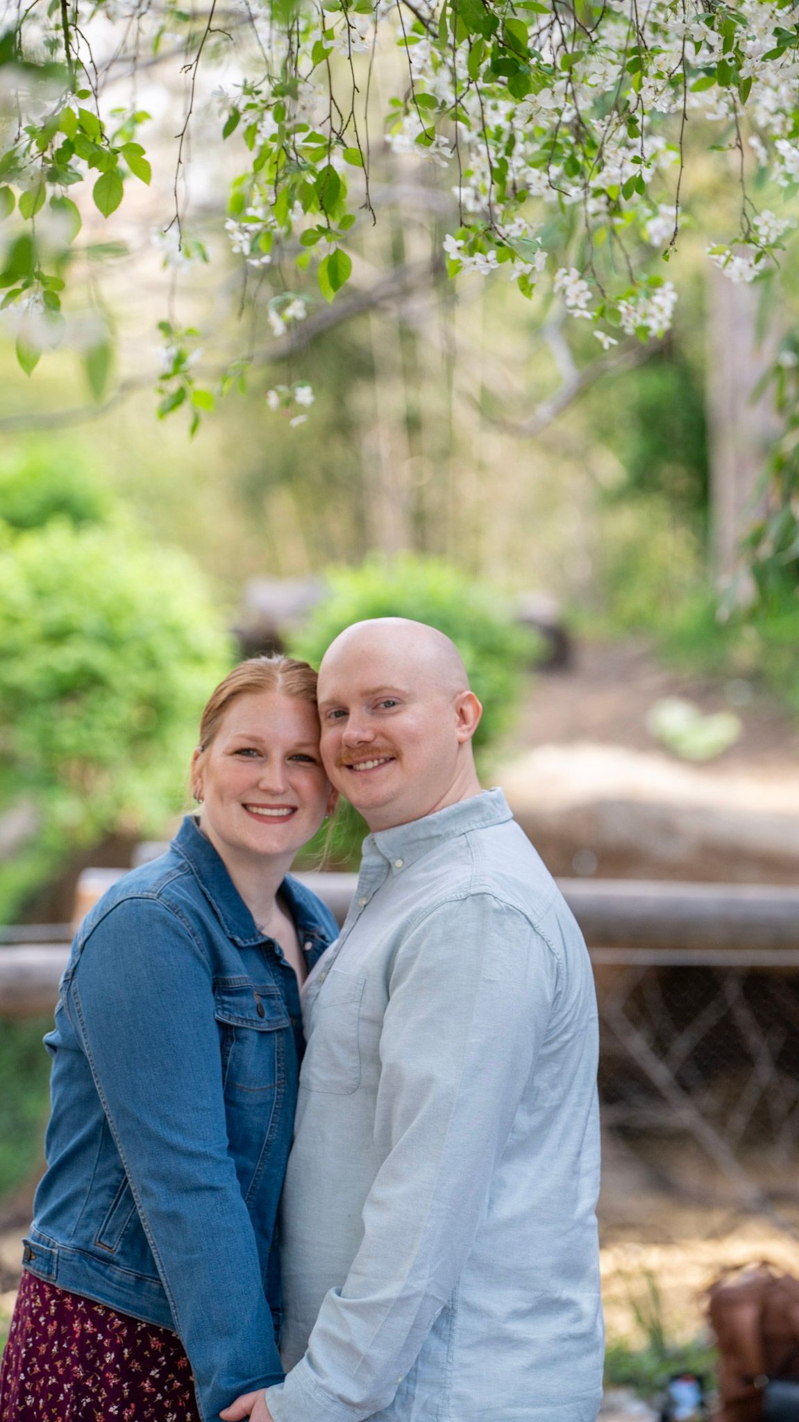 A couple poses together under blooming spring tree branches in a garden setting.