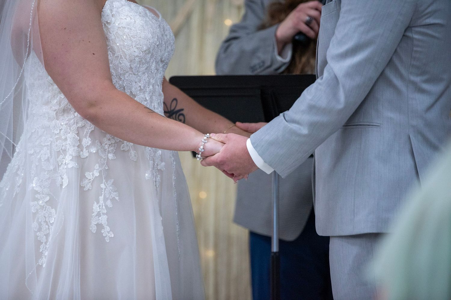 Close-up of joined hands during a wedding ceremony showing lace dress details and light gray suit.