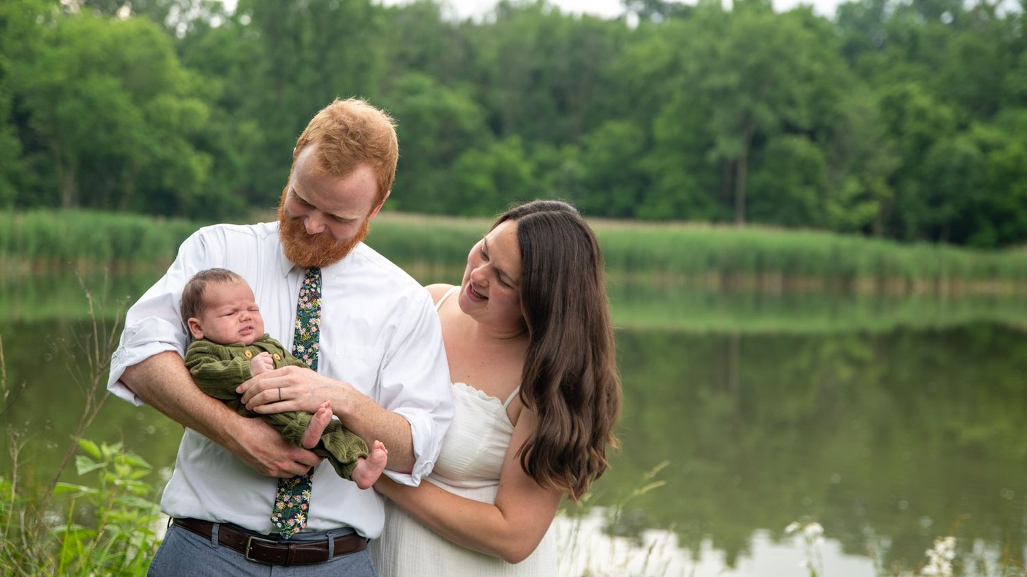 Parents share loving smiles while holding their newborn baby outdoors by a peaceful lake.
