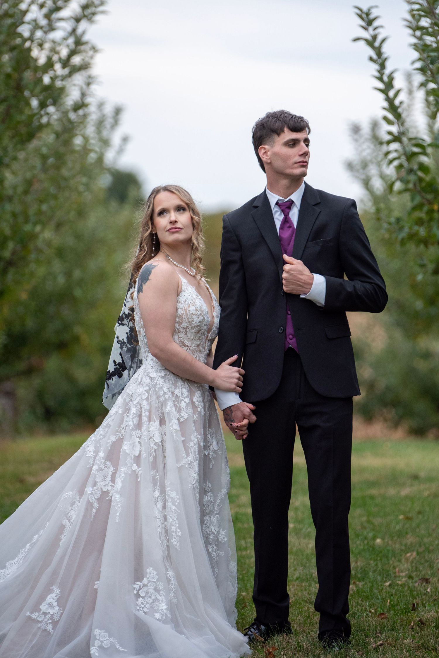 A couple stands together in an orchard wearing wedding attire during an autumn photoshoot.