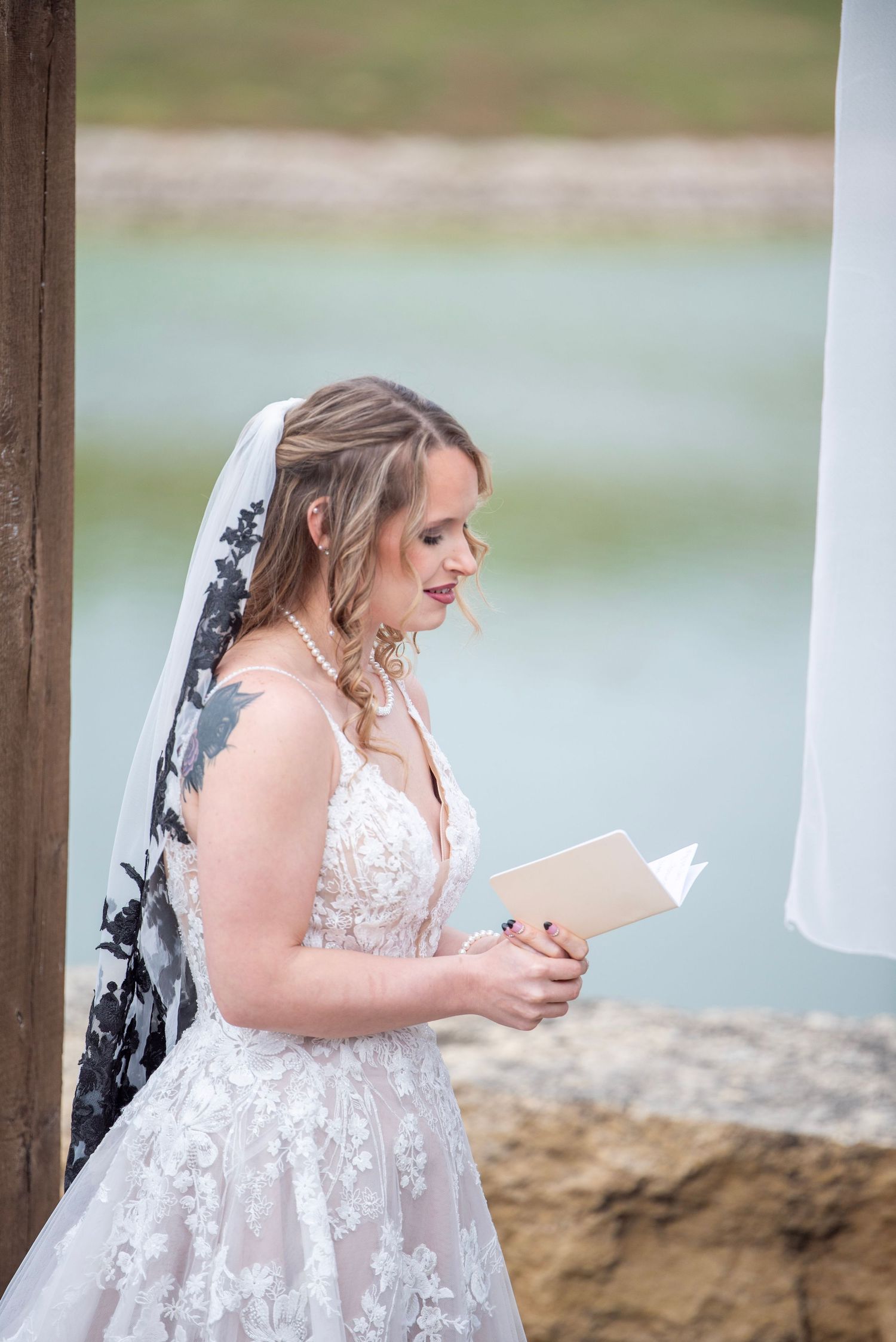 Bride in lace dress reading from paper during lakeside ceremony.