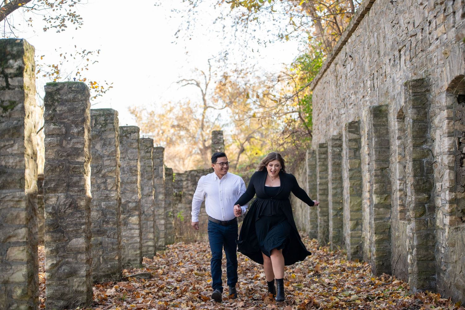 A couple walks together between stone columns on a path covered in fall foliage.