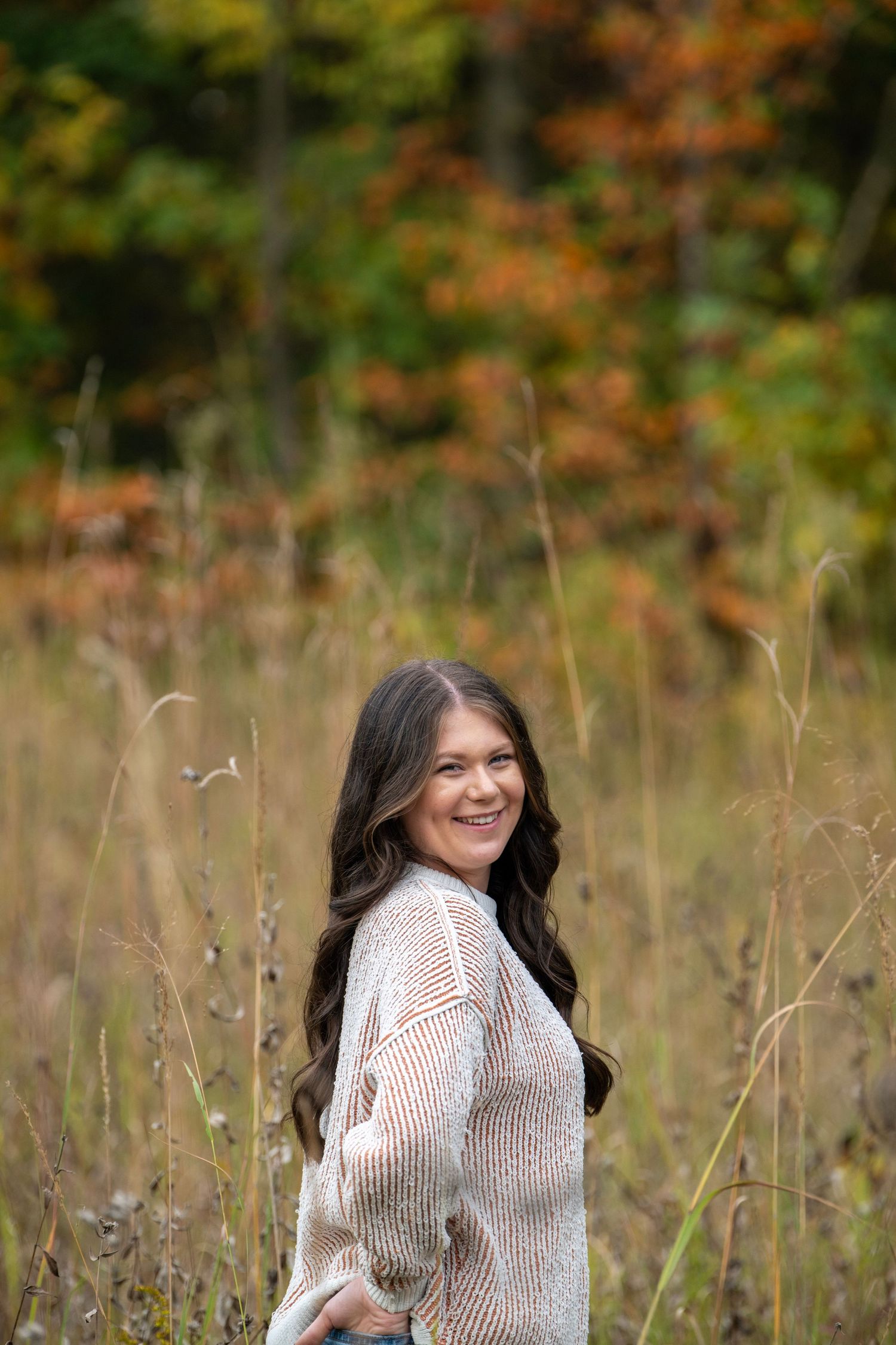 A person in a cream sweater poses in an autumn field with golden foliage in the background.