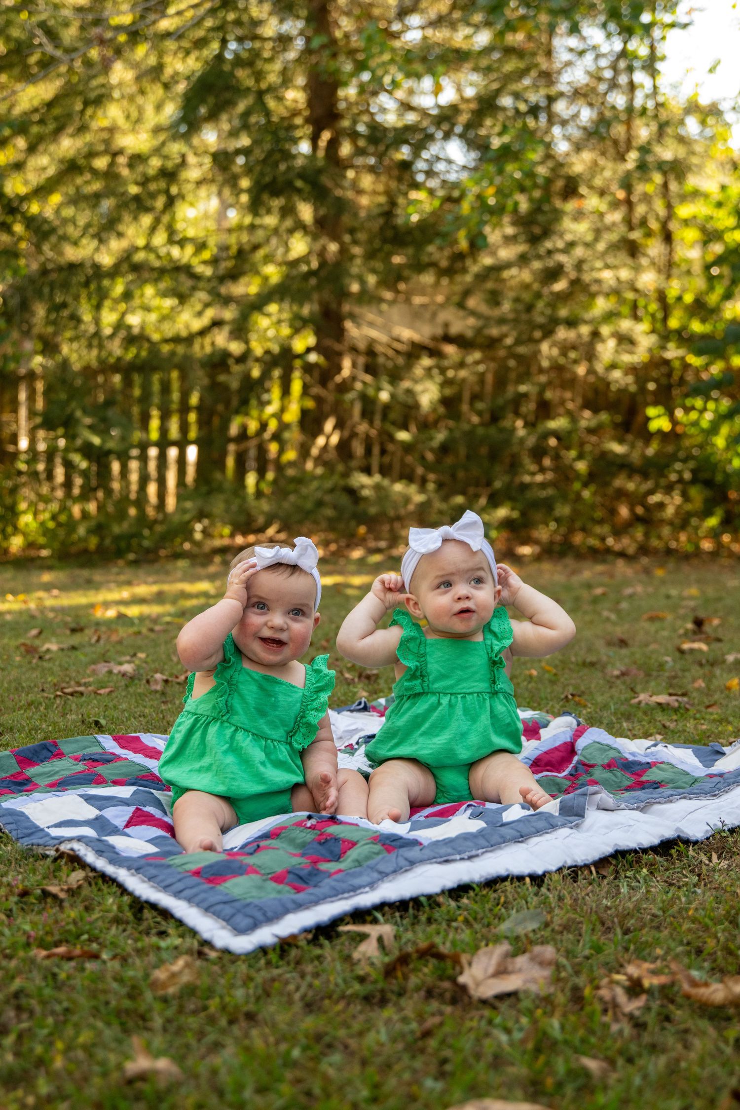 Baby in bright green dress sitting on colorful patchwork blanket outdoors with sunlit trees in background.